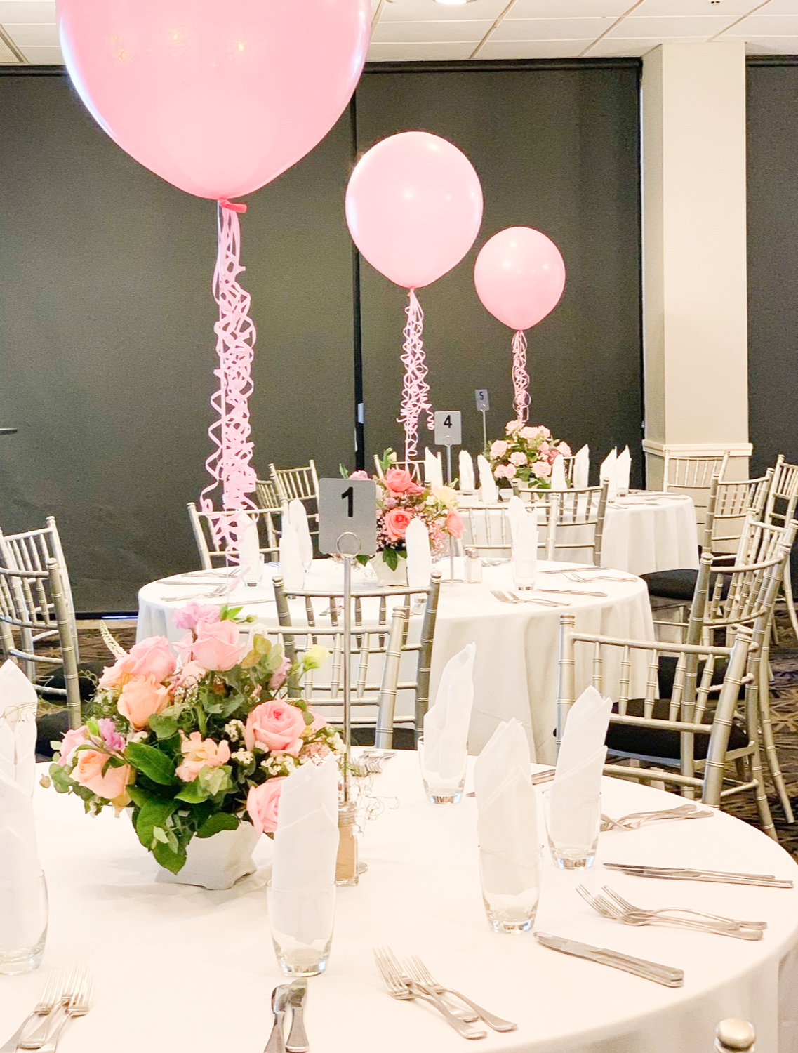 Elegant event dining setup with round tables covered with white tablecloths, pink balloon centerpieces with curly ribbons, floral arrangements with pink roses and greenery, white napkins, and silverware, in a decorated banquet hall.