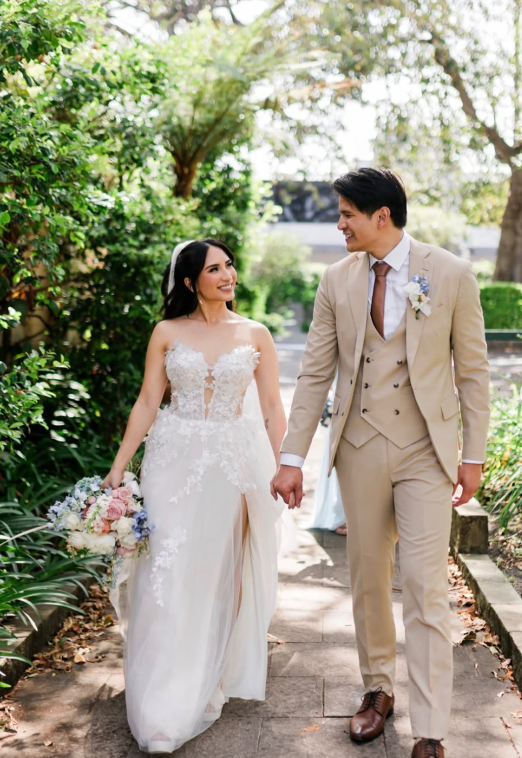 A bride and groom walk hand in hand outdoors on a sunny day, smiling at each other. The bride wears a white lace wedding gown, and the groom wears a beige suit with a brown tie. They are surrounded by greenery and trees.