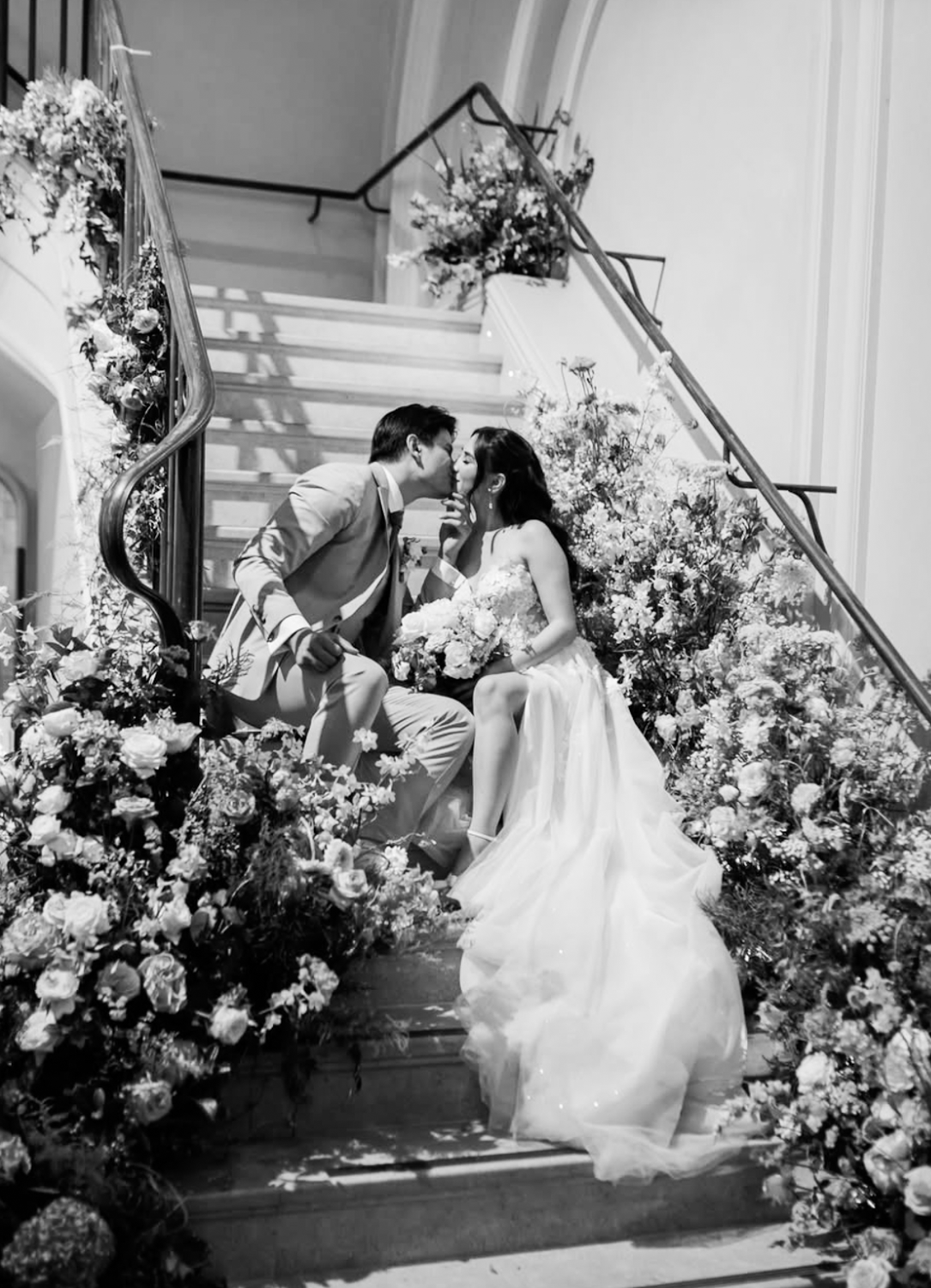 A black and white photograph of a bride and groom sitting on a staircase decorated with flowers, sharing a kiss at their wedding.