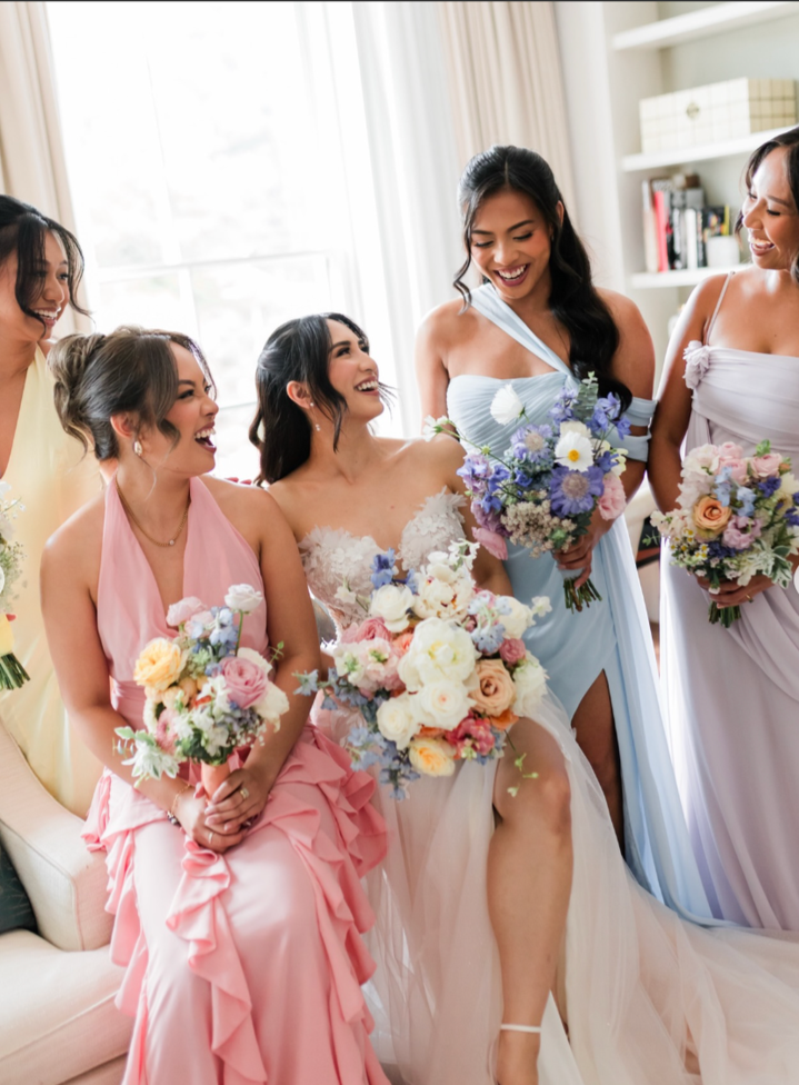 A group of five women, including a bride in a white wedding dress, smiling and holding bouquets of flowers, standing in a bright room with a window and bookshelf in the background.