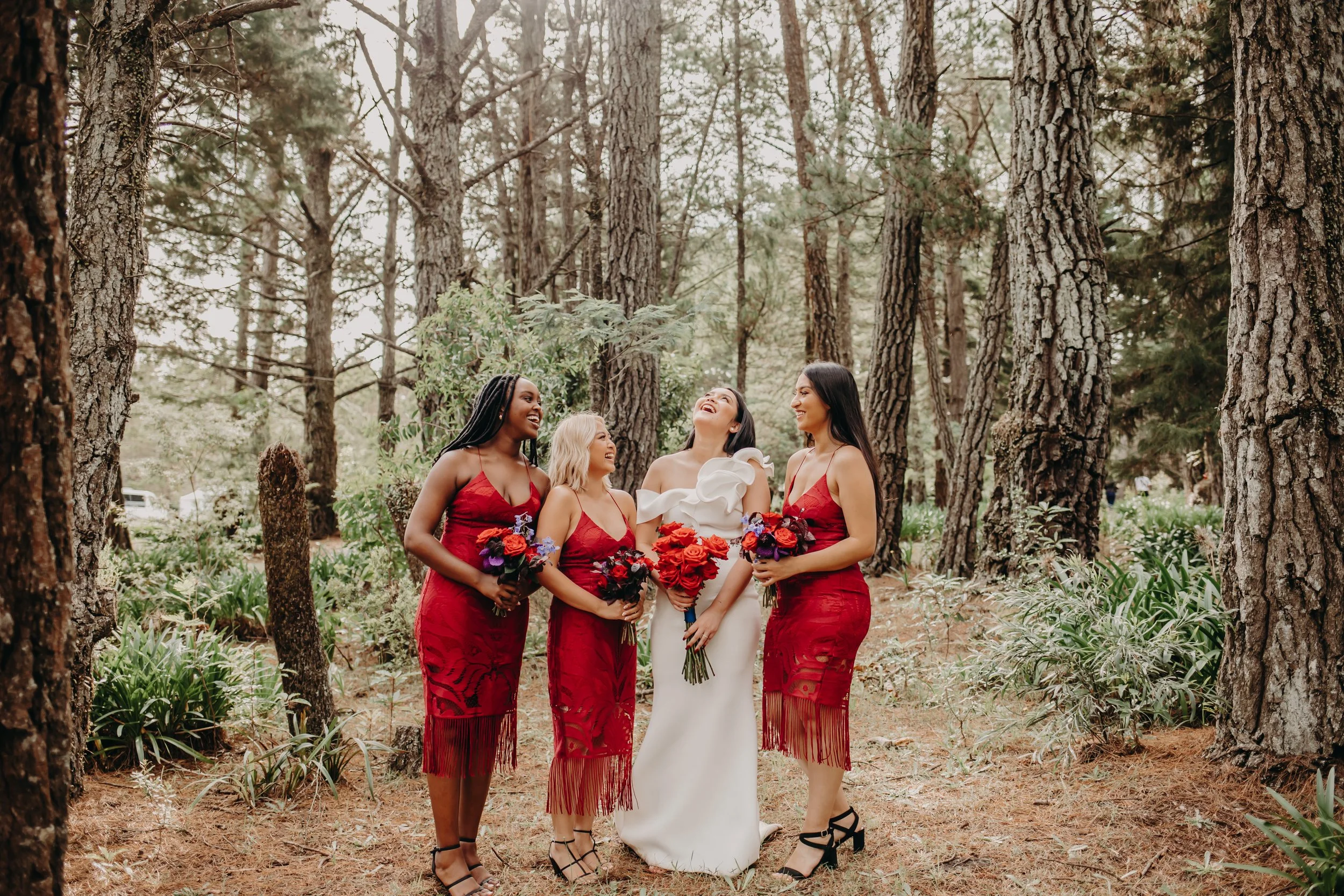 Group of five women in dresses standing in a forest, sharing a laugh while holding flower bouquets, with trees and greenery in the background.