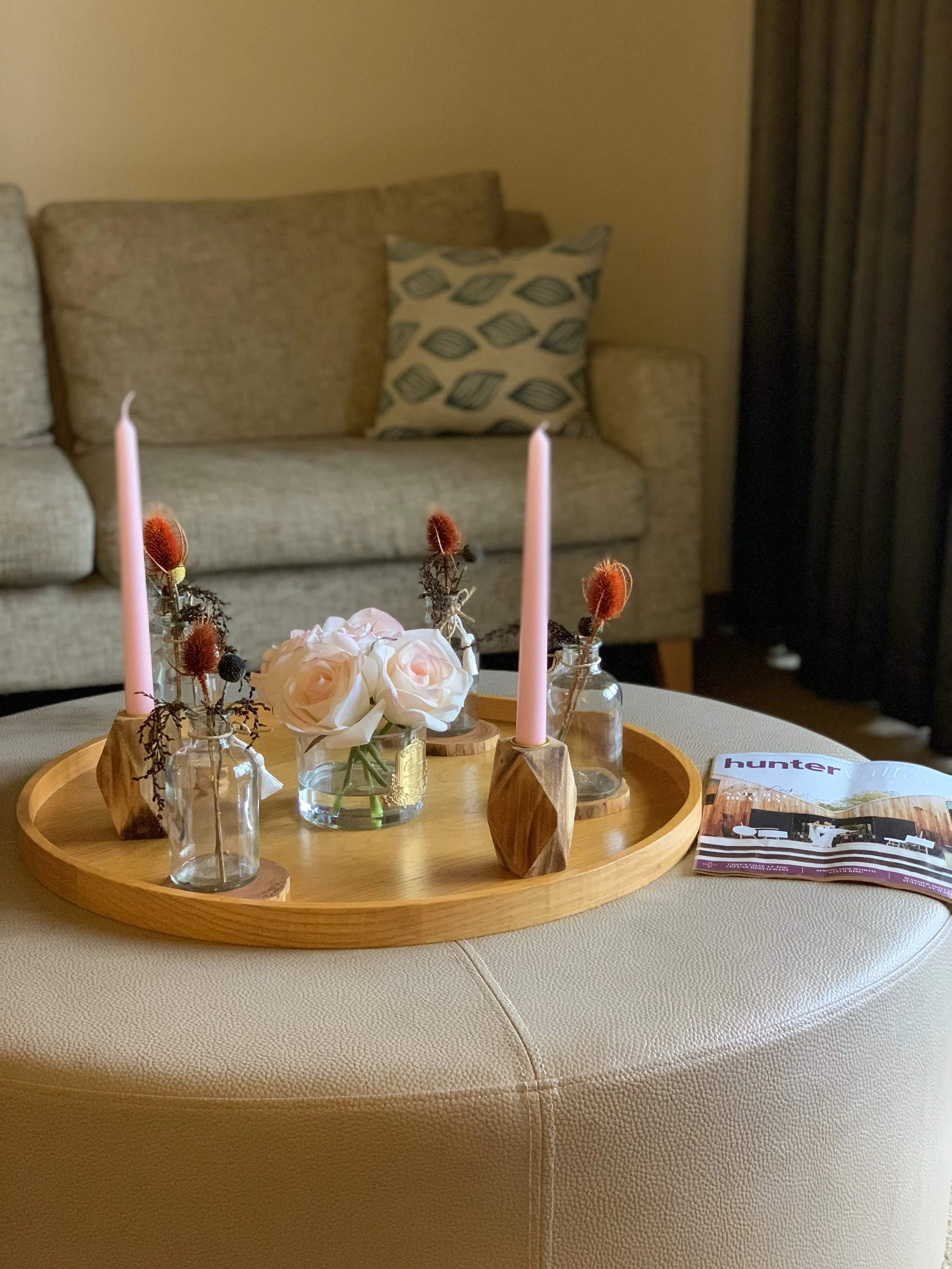 A wooden tray with glass vases holding white roses and dried flowers, two pink taper candles, and a magazine on a beige upholstered surface in a living room setting.