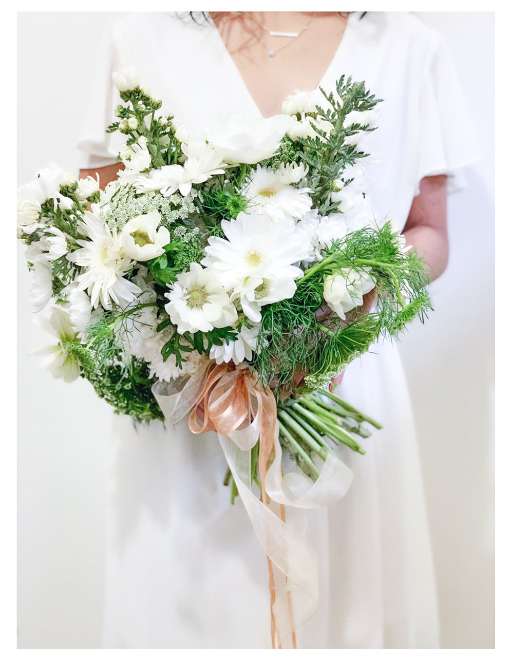 Person holding a bouquet of white flowers and greenery, wrapped with ribbons, against a white background.