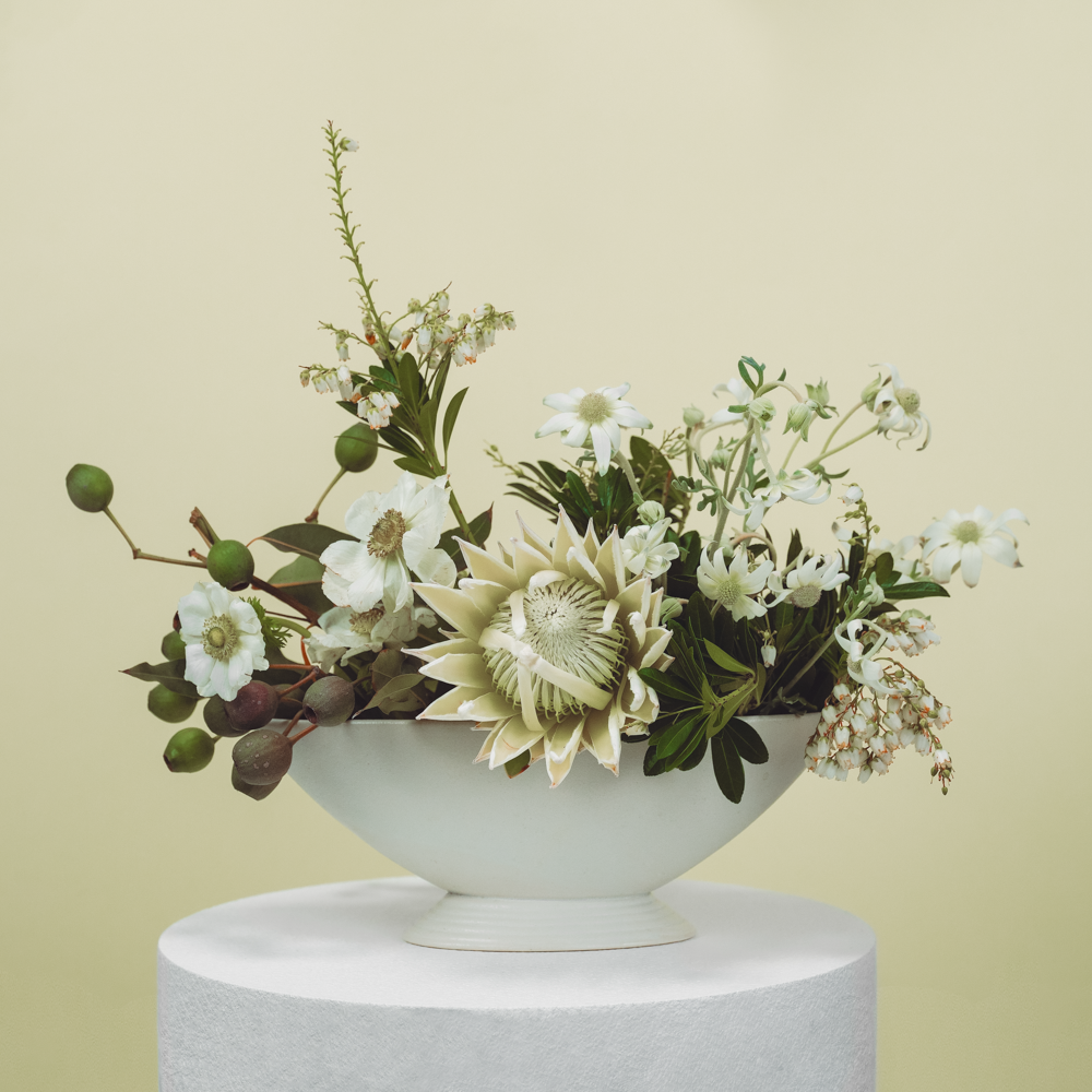 A white ceramic bowl with a variety of white flowers and greenery, placed on a white pedestal against a plain, light background.