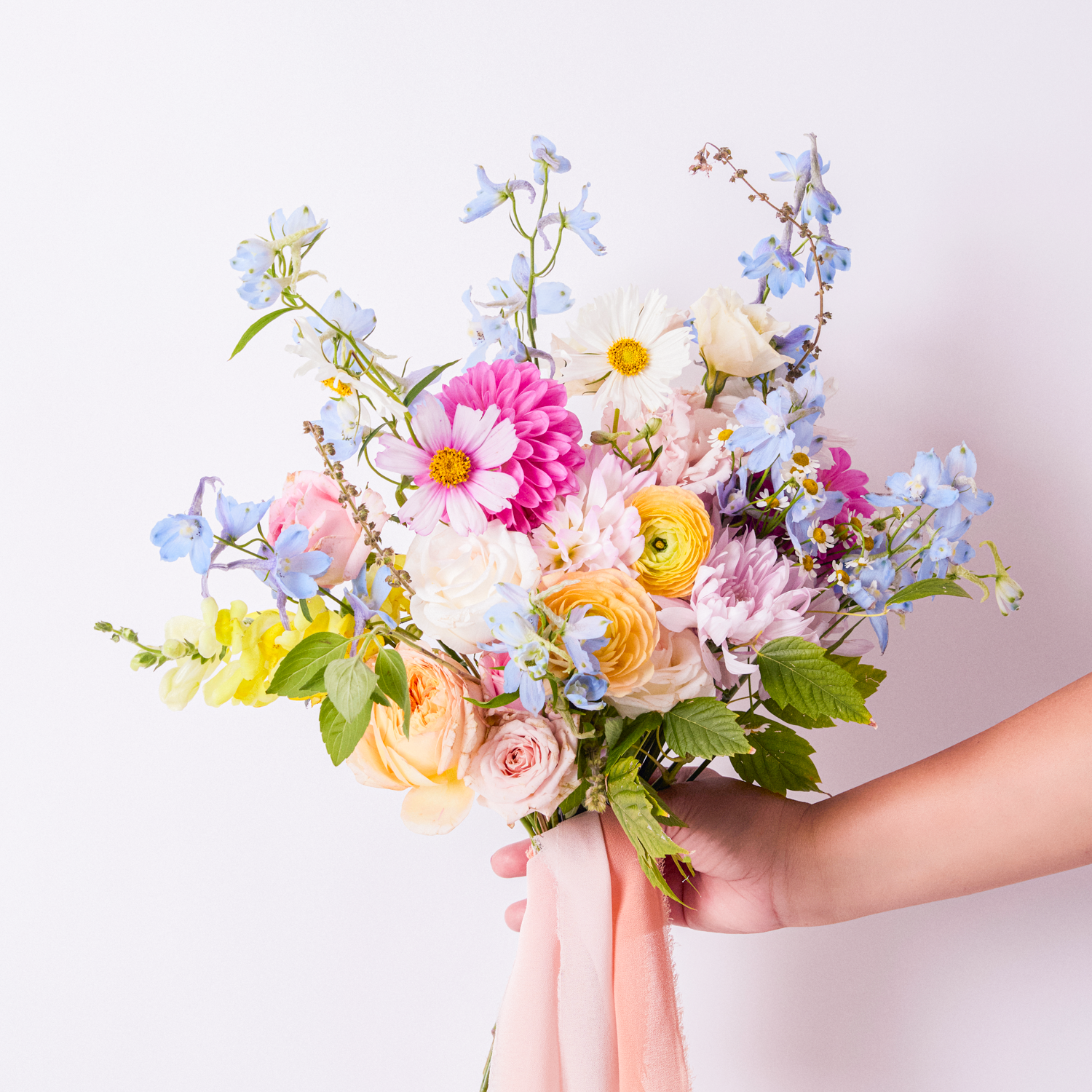 A person holding a colorful bouquet of mixed flowers, including pink, white, yellow, purple, and blue blossoms, tied with a pink ribbon, against a white background.