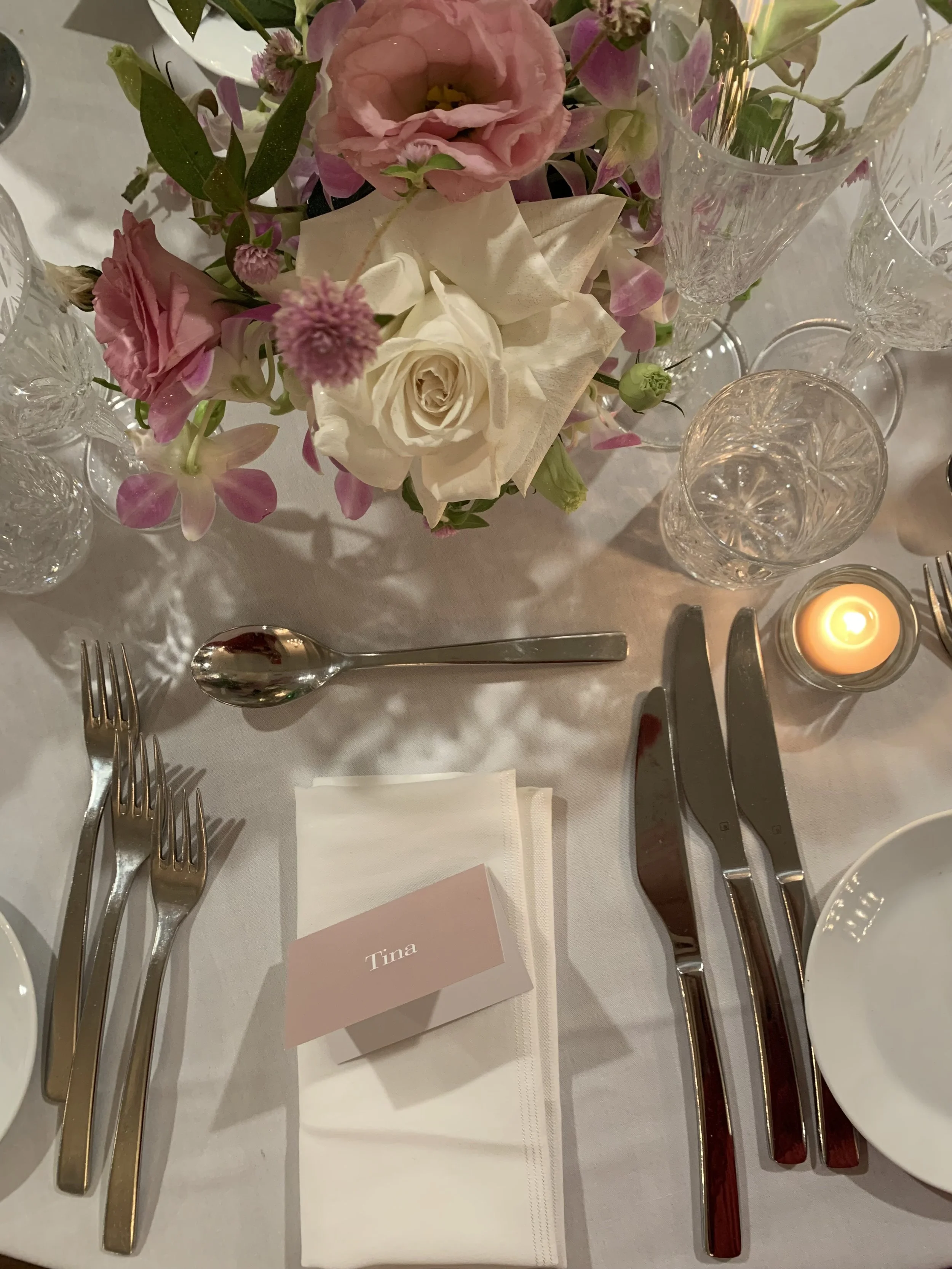 A formal table setting with silverware, crystal glasses, a lit candle, a floral centerpiece with pink and white flowers, a place card with the name 'Tina', and a white napkin.