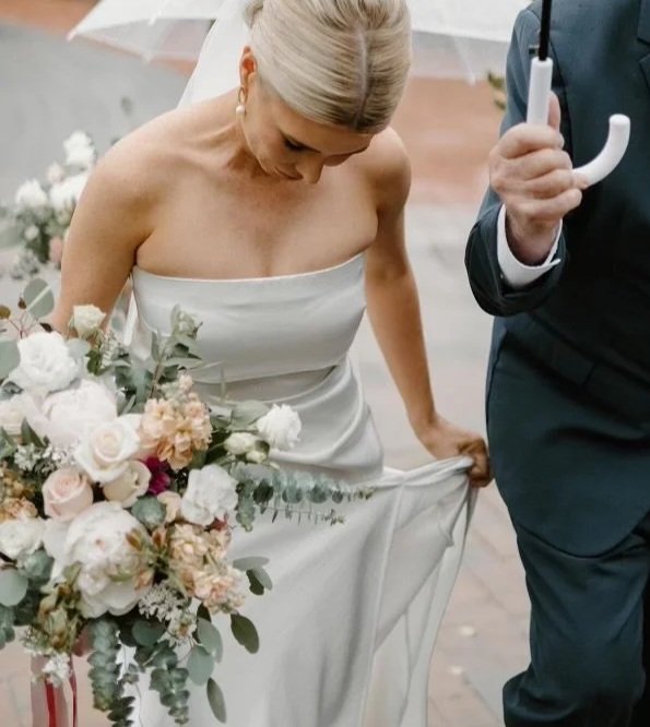 Bride holding her wedding dress while walking, with a bouquet of flowers, next to a man holding an umbrella.