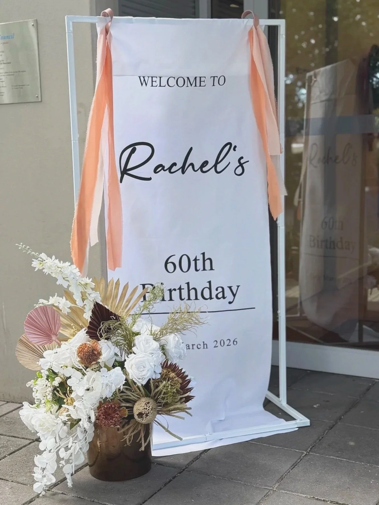 A white fabric banner with black text that reads, "Welcome to Rachel's 60th Birthday." The banner is decorated with soft peach ribbons on each side, and there is a large bouquet of white and brown flowers in a vase placed in front of it. The event ap