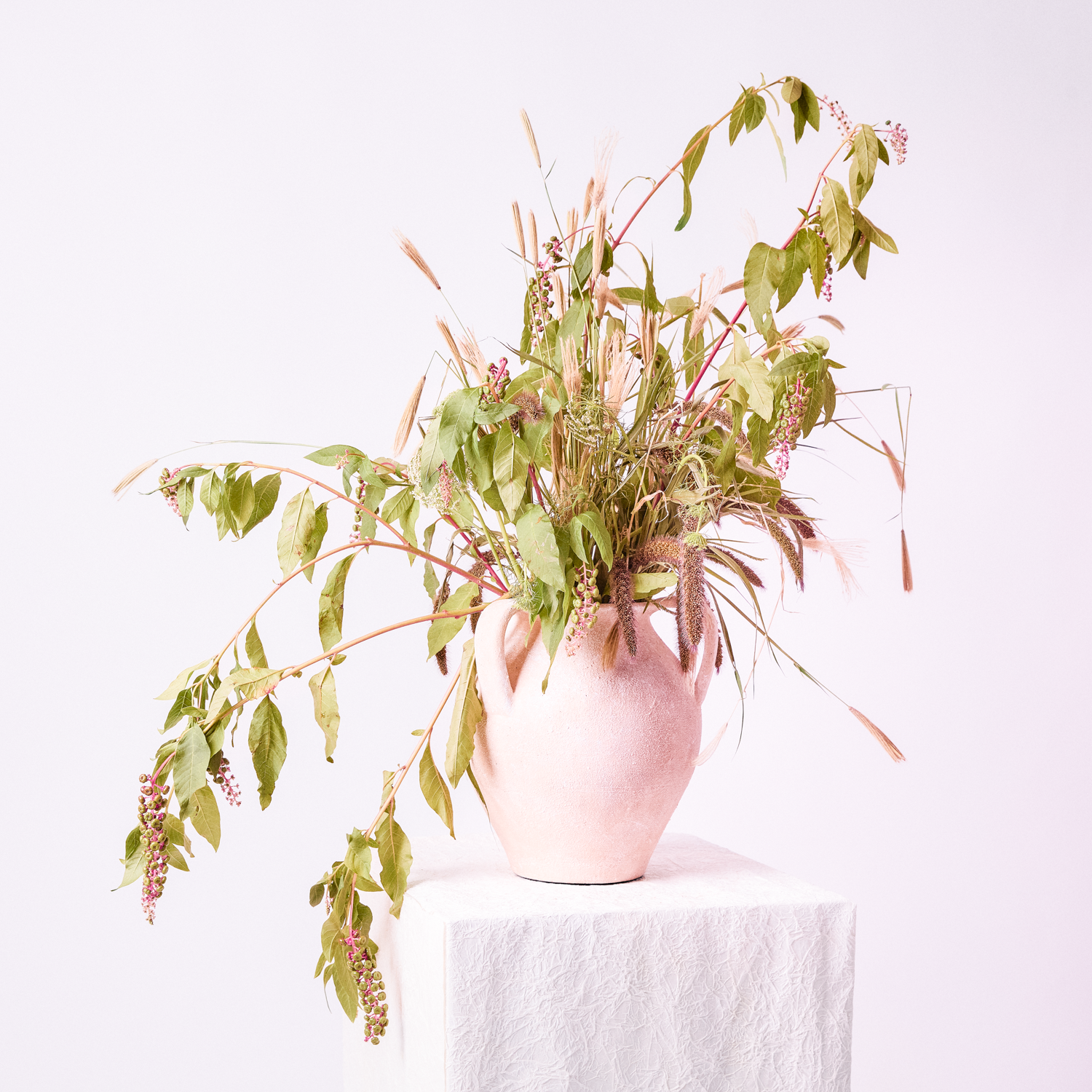 A pink ceramic vase filled with a variety of dried and artificial plants and flowers on a white textured pedestal against a plain white background.