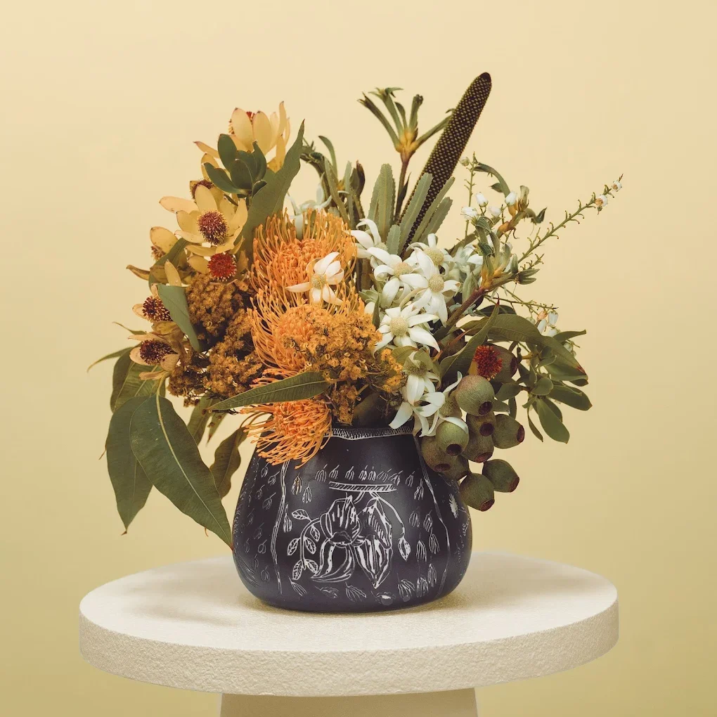 Flower bouquet with various flowers and greenery in a black vase on a white pedestal against a beige background.