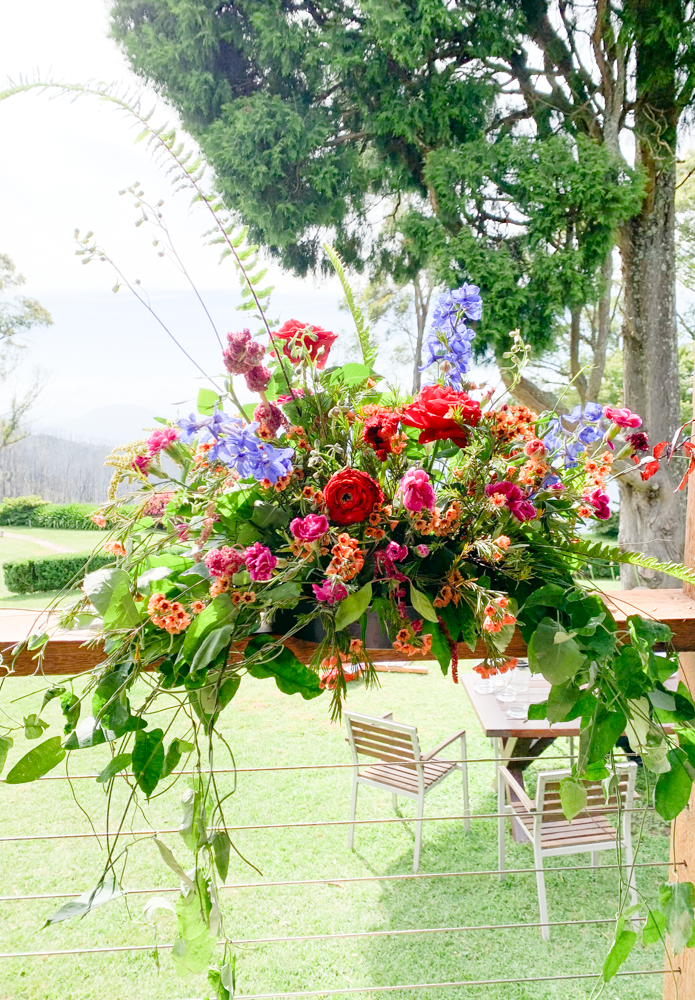 Colorful flower arrangement with pink, red, purple, and orange blooms on a balcony railing, overlooking a lush green yard with trees and outdoor furniture.