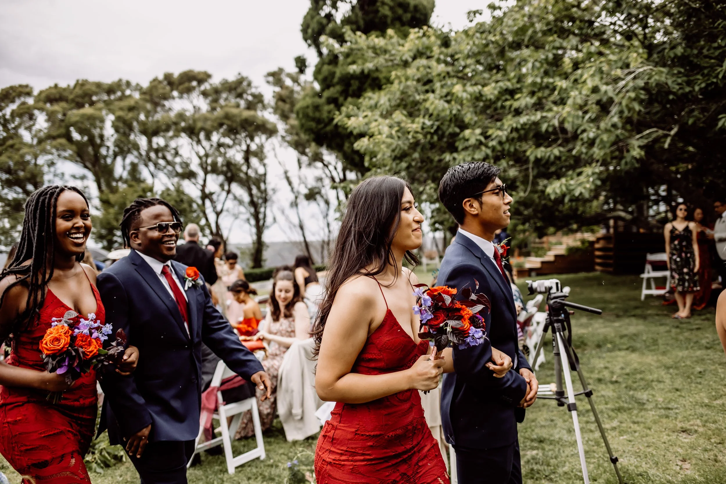 A group of people in formal attire, including women in red dresses and men in suits, participate in a wedding ceremony outdoors on a cloudy day, holding bouquets of flowers.