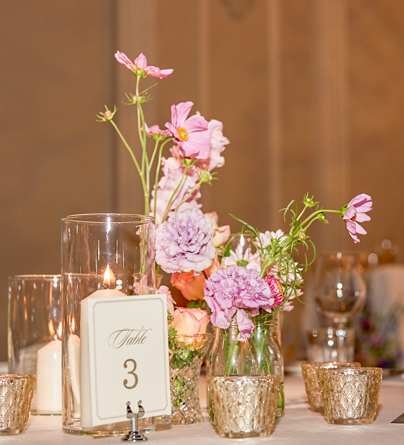 A table centerpiece with pink, purple, and white flowers in glass vases, with candles and a table number card labeled 'Table 3' in a glass holder.
