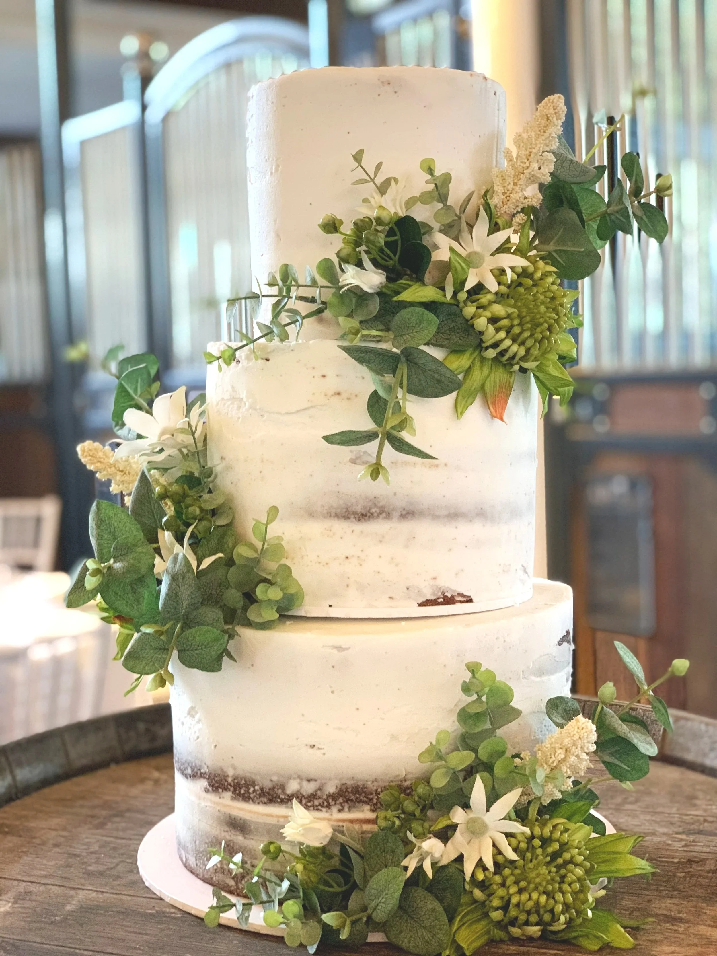 A two-tier white wedding cake decorated with green leaves, white flowers, and green and white berries, sitting on a wooden table.