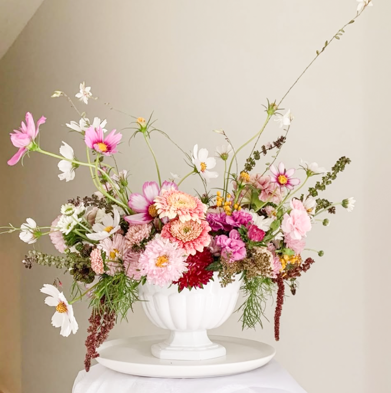 A white vase filled with pink, white, and red flowers, including cosmos, dahlias, and other blooms, sitting on a white table against a beige wall.