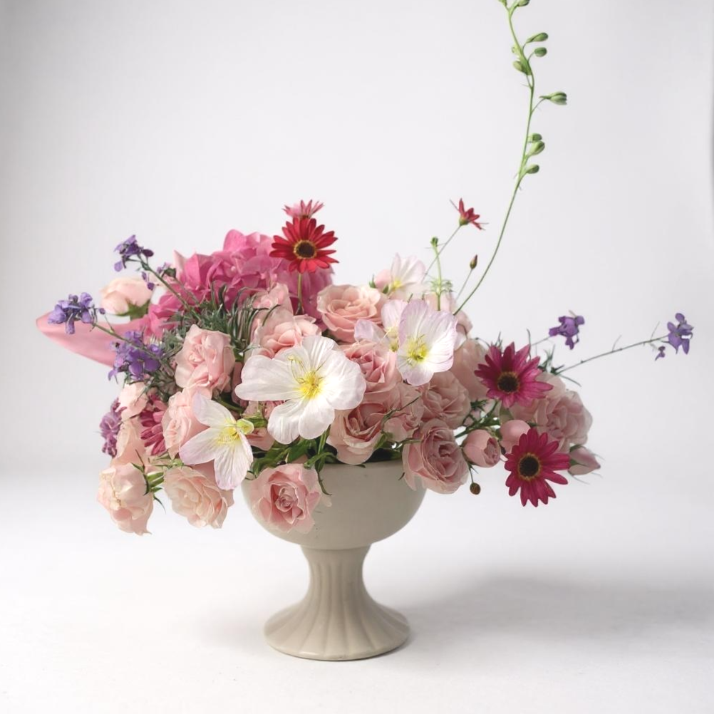 A white vase filled with pink, purple, and white flowers, arranged against a plain white background.