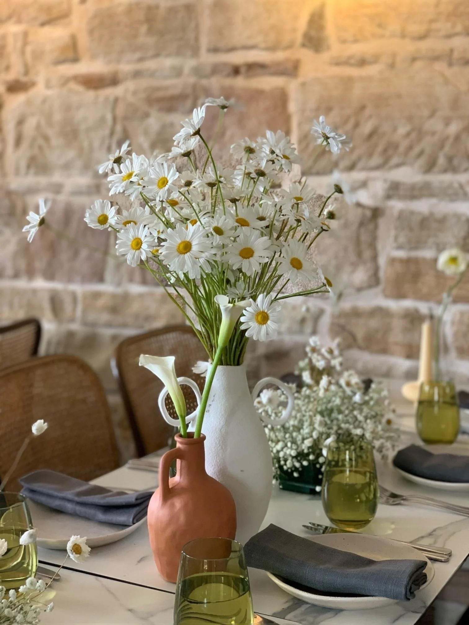 A dining table decorated with a bouquet of white daisies in a white vase, surrounded by green glasses, gray napkins, and dishes, with a brick wall background.