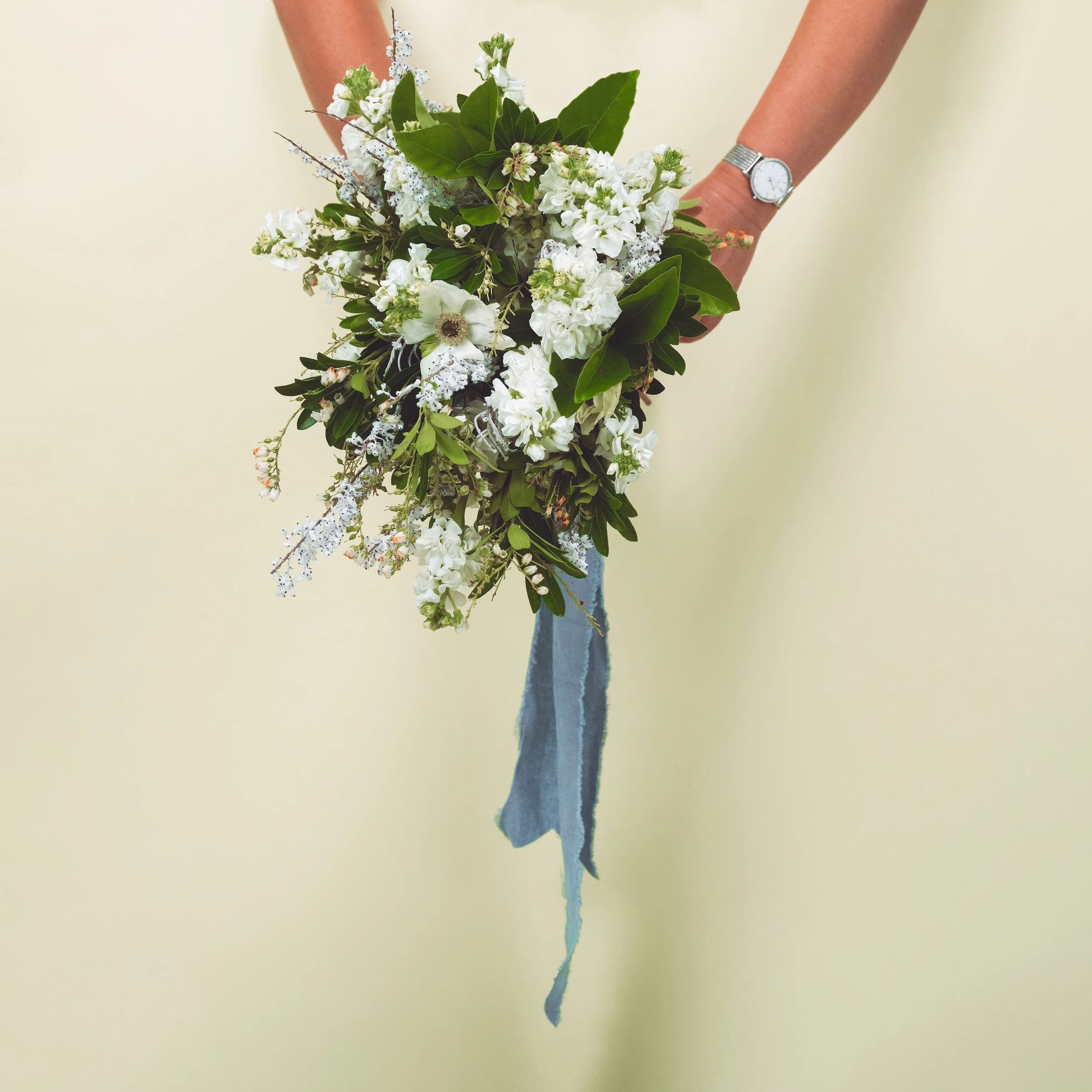 Two hands holding a bouquet of anemones spring foliage and lily of the valley tied with slate blue linen ribbon