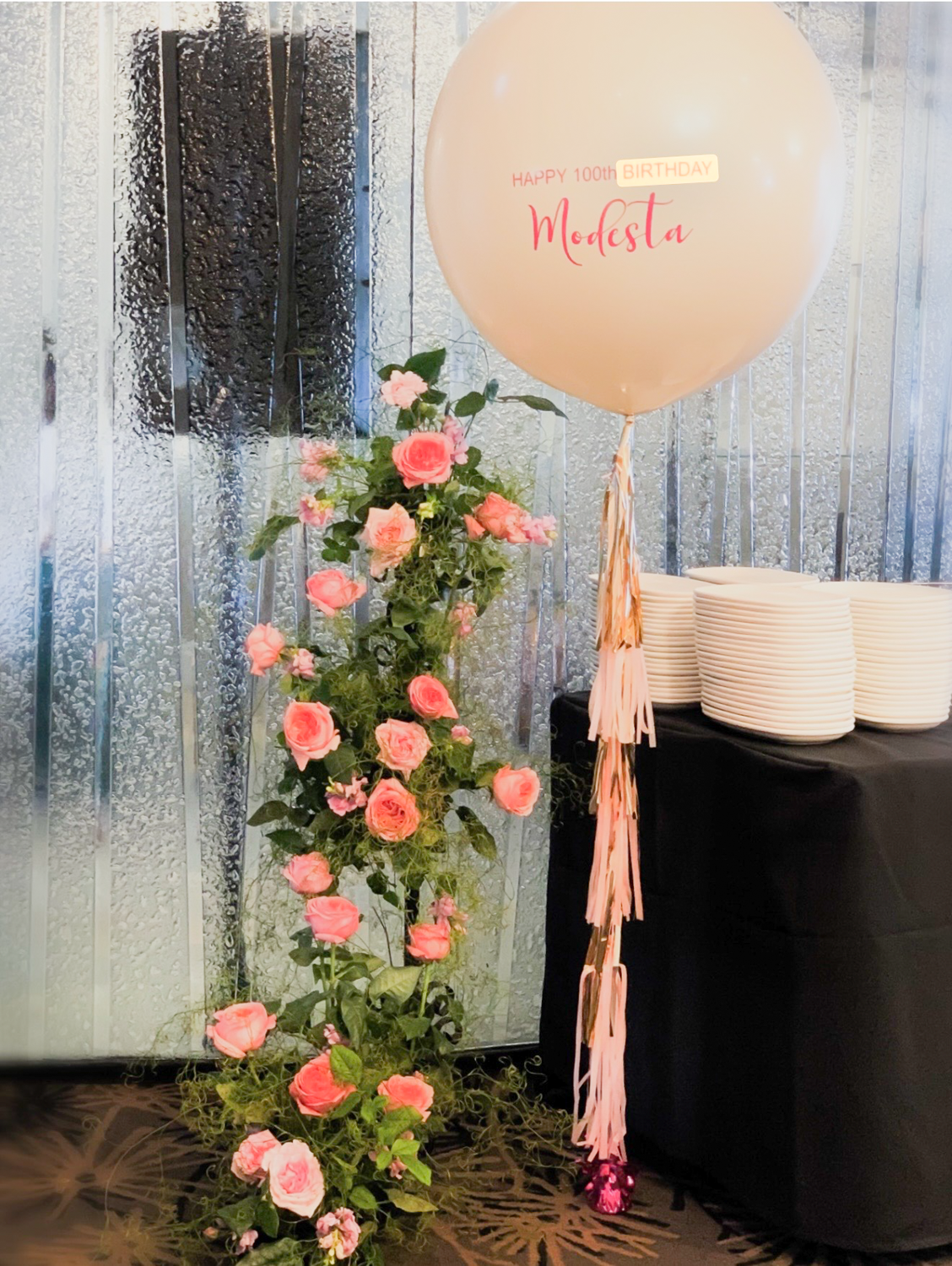 Pink roses climbing a green trellis next to a white balloon with pink and yellow text that says "HAPPY 100th BIRTHDAY Modesta". There are stacks of white plates on a table covered with a black cloth, and the background features a metallic textured wa