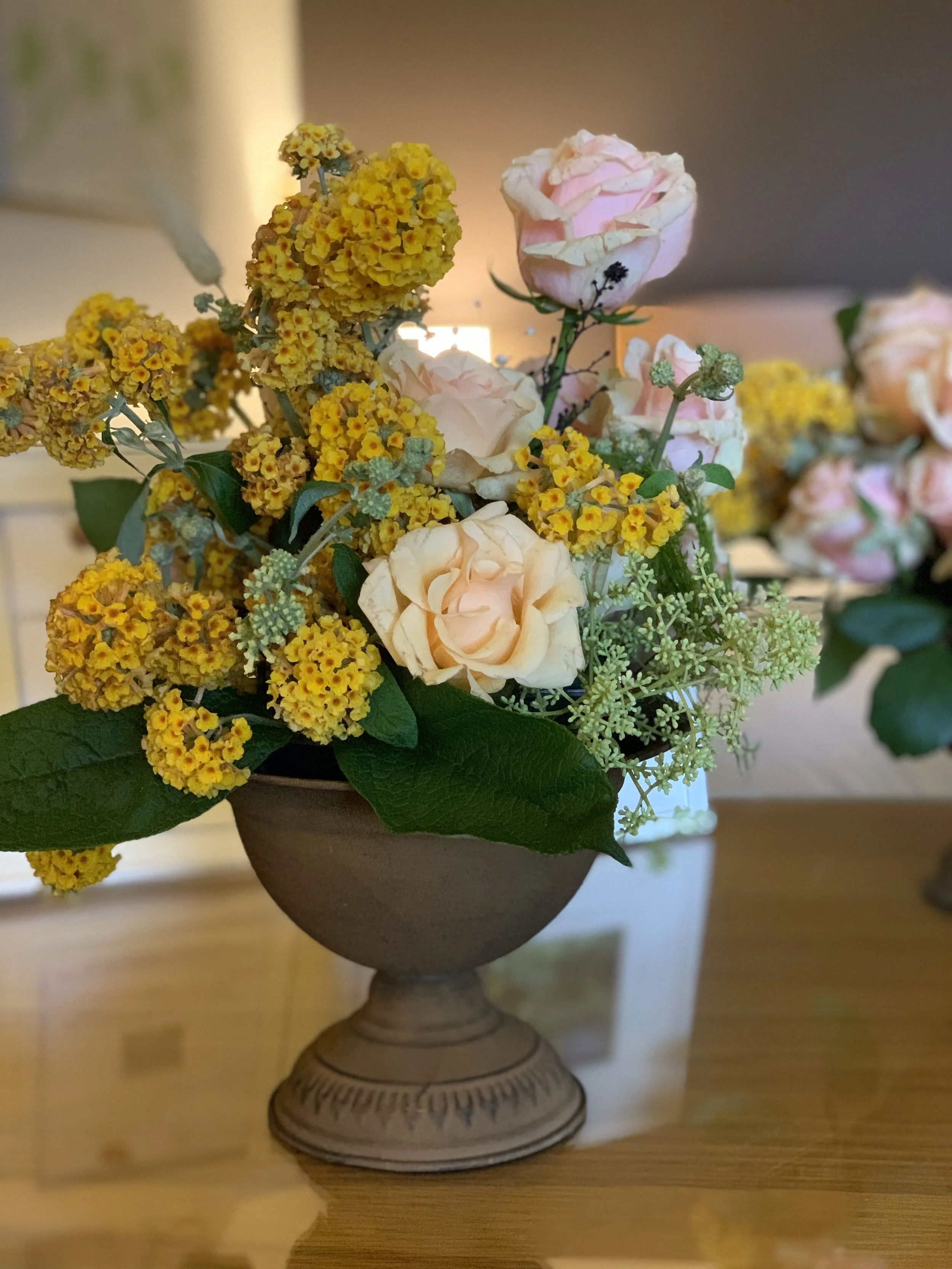 A floral arrangement featuring light pink roses, yellow flowers, and green leaves in an ornate gray pedestal vase on a wooden surface.