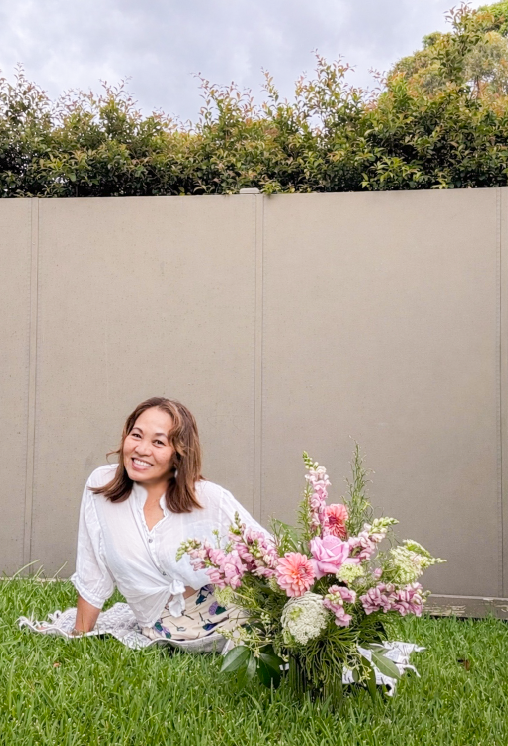 A woman with shoulder-length brown hair, wearing a white shirt, is sitting on the grass next to a large bouquet of pink and white flowers in an outdoor setting, with a beige fence and green foliage in the background.