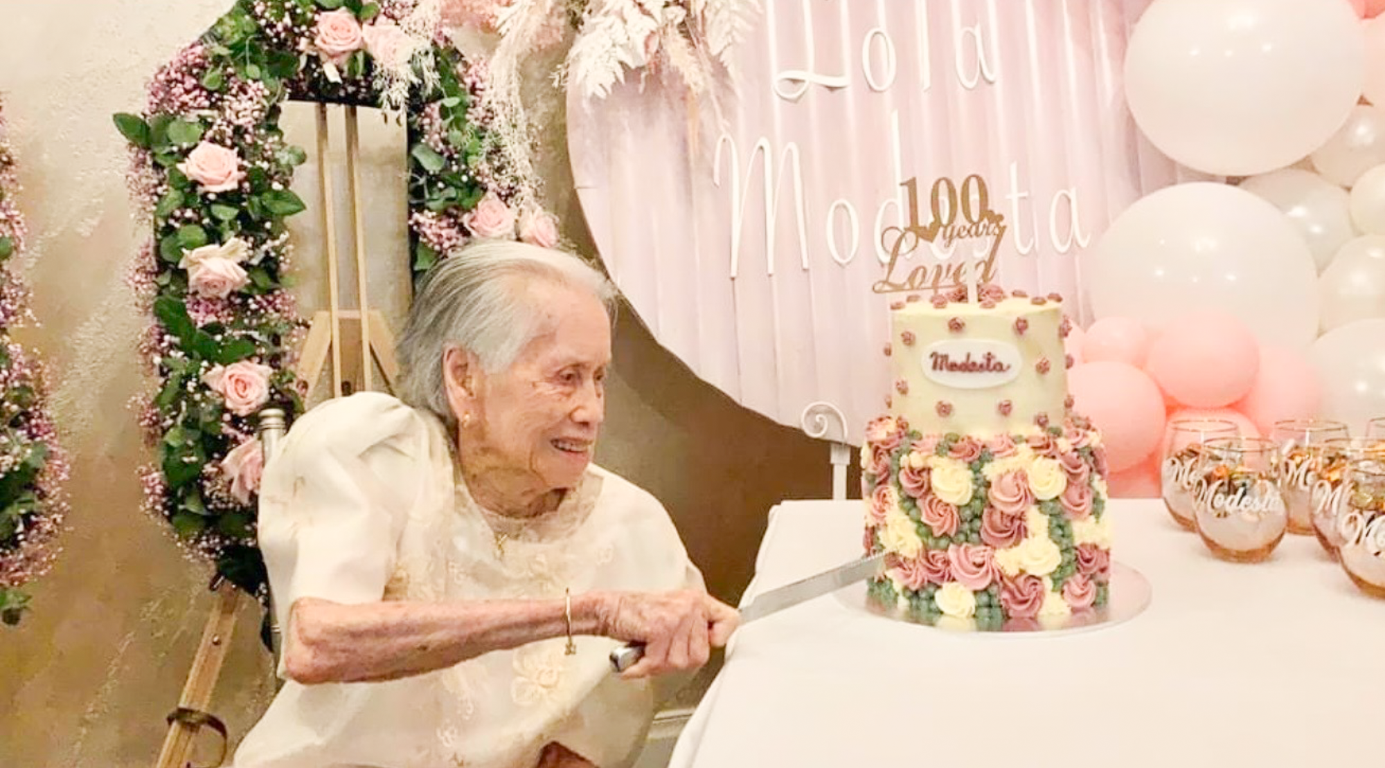 An elderly woman with gray hair and a white dress is cutting a birthday cake decorated with pink, yellow, and purple roses at a celebration. The background features pink balloons, floral decorations, and a sign with partially visible words.