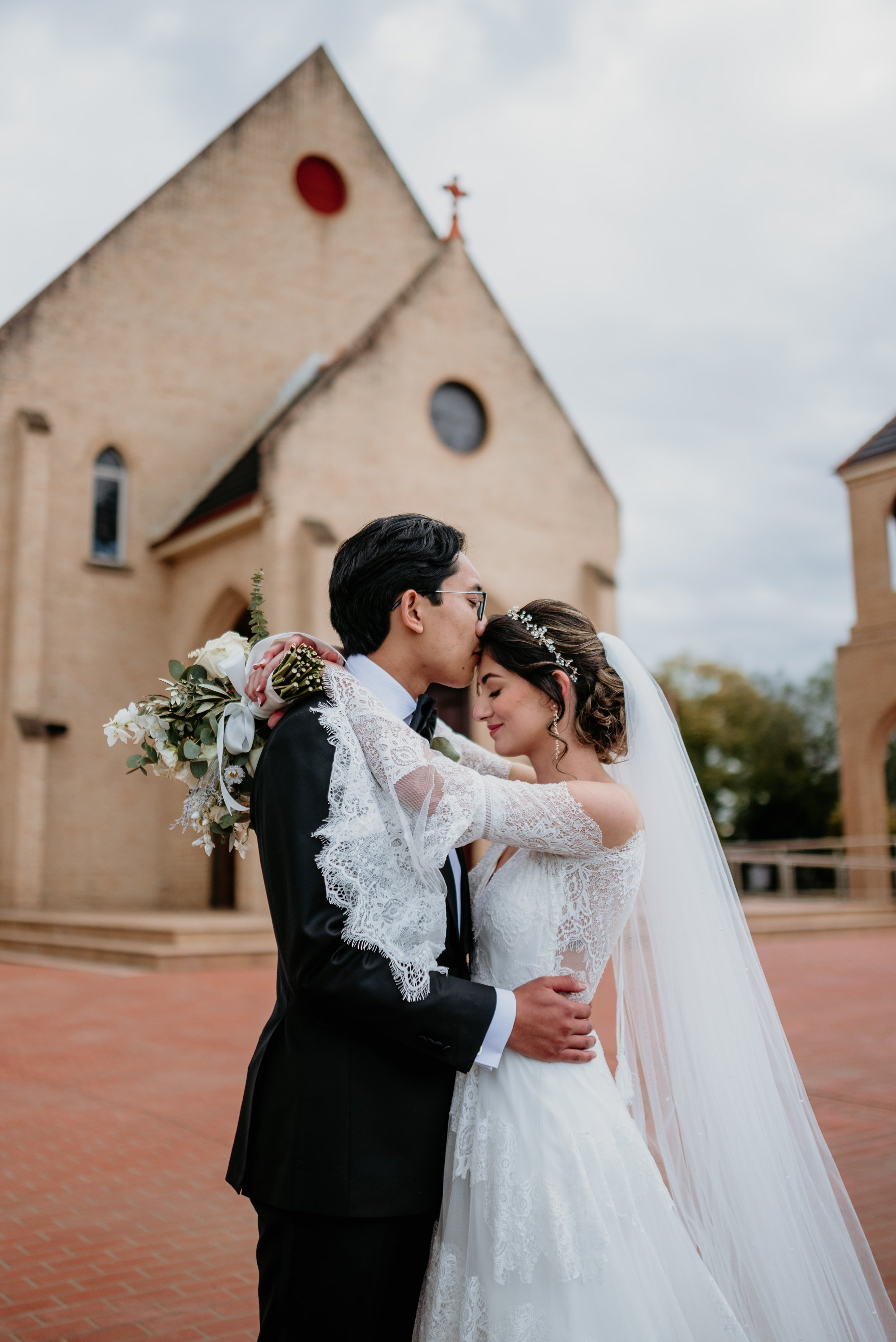 A bride and groom embracing and kissing outdoors in front of a church, with a cloudy sky overhead.