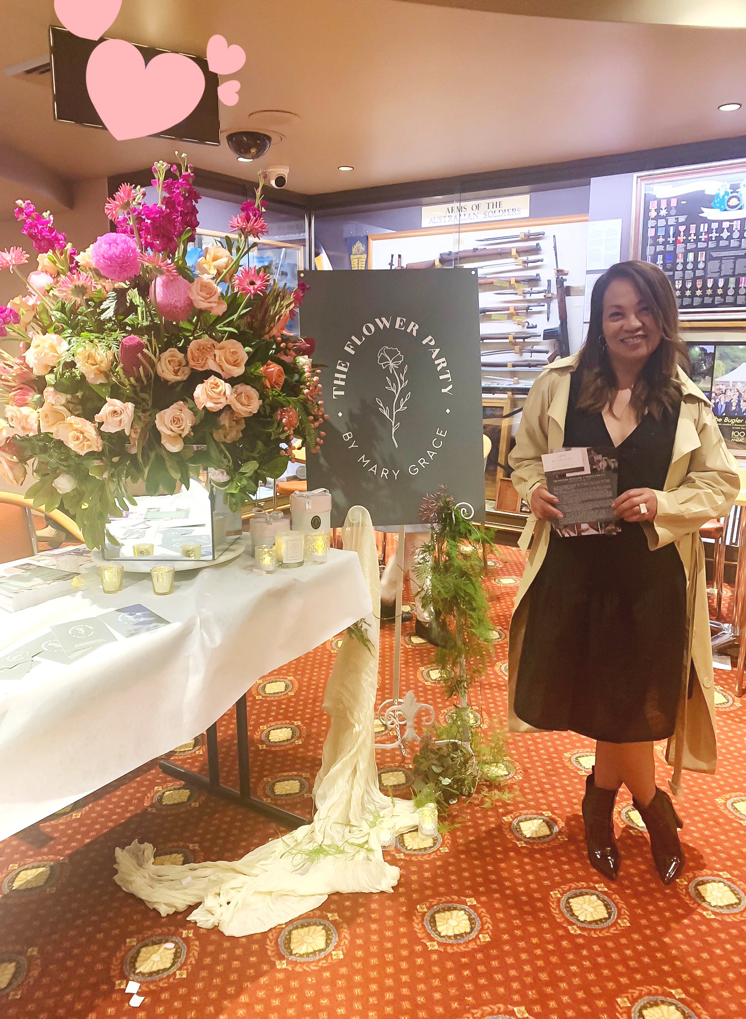 A woman standing next to a table decorated with a large floral arrangement, candles, and brochures at the 'The Flower Party' event, with display cases and a sign in the background.