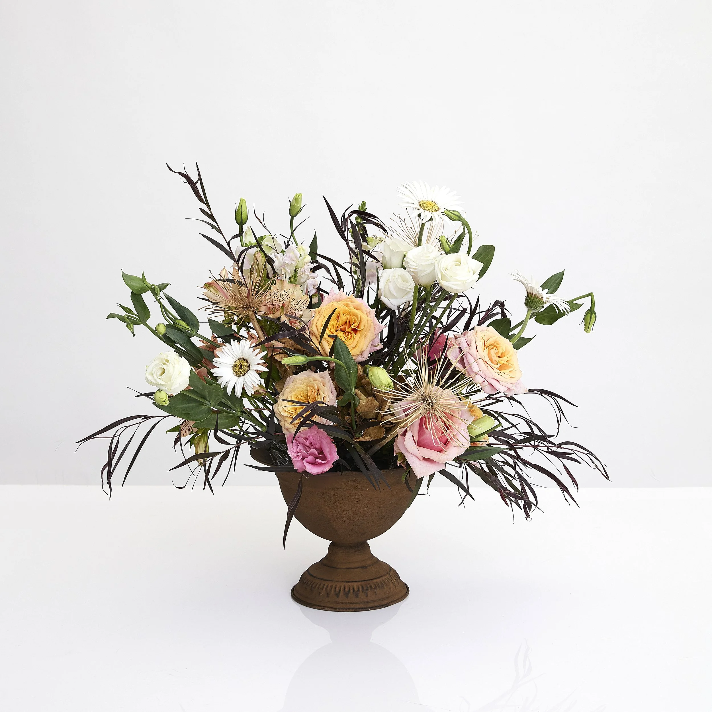 A floral arrangement in a brown pedestal vase featuring roses, daisies, and other flowers against a plain white background.