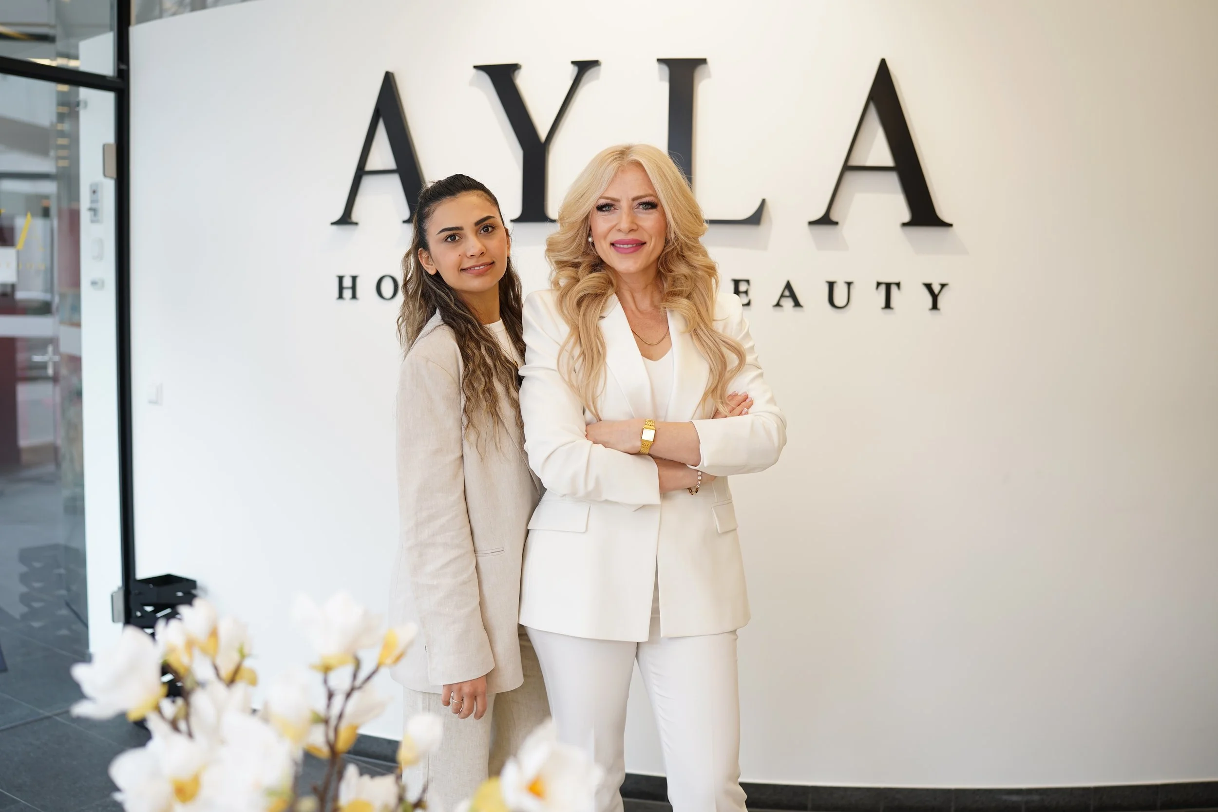 Zwei Frauen stehen vor einer weißen Wand mit großen schwarzen Buchstaben, die "AYLA HOUSE OF BEAUTY" sagen.