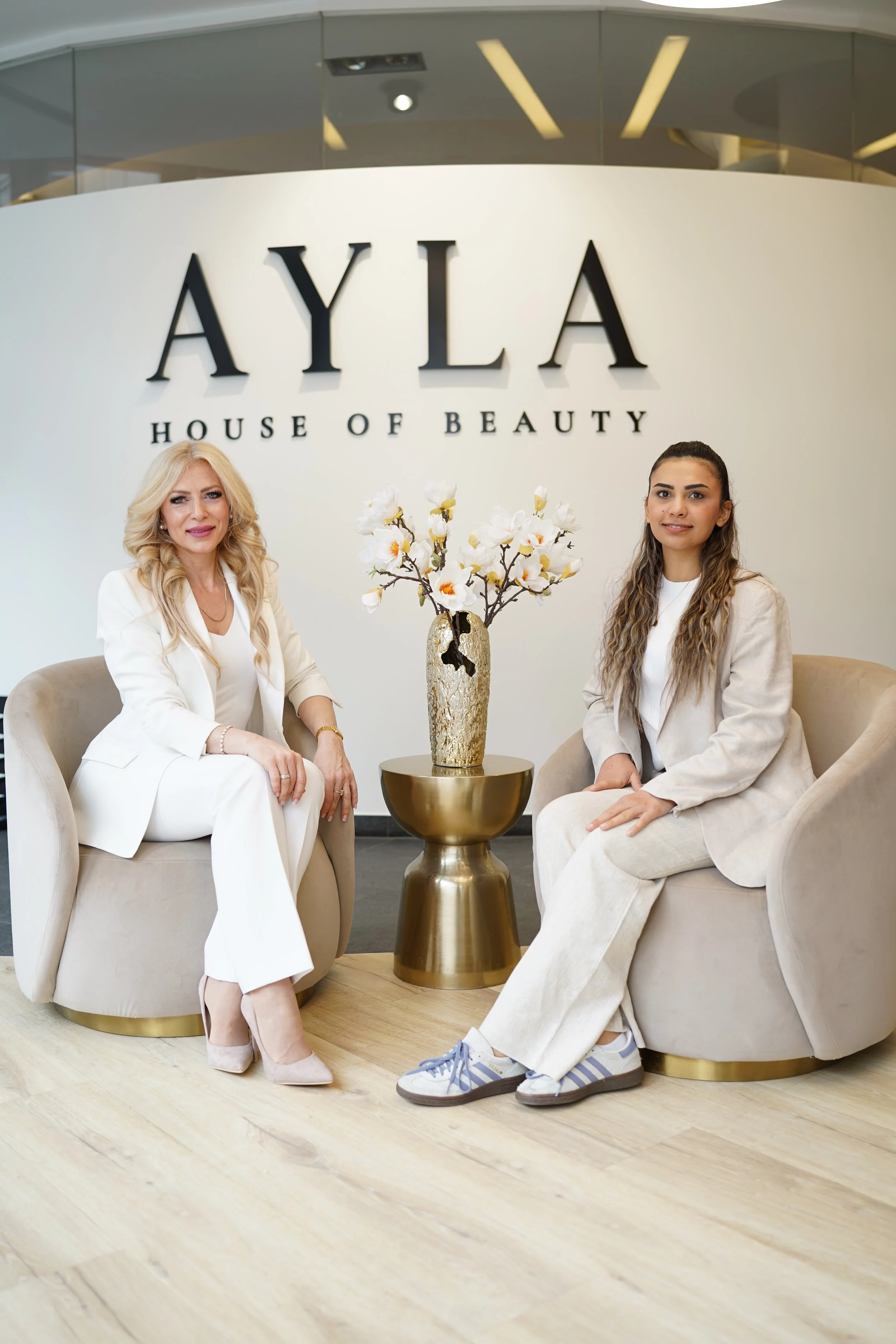 Zwei Frauen sitzen in einem Schönheitsstudio namens 'AYLA House of Beauty' auf beigen Sesseln, mit einem goldfarbenen Tisch mit einer dekorativen Vase voller weißer Blumen dazwischen.