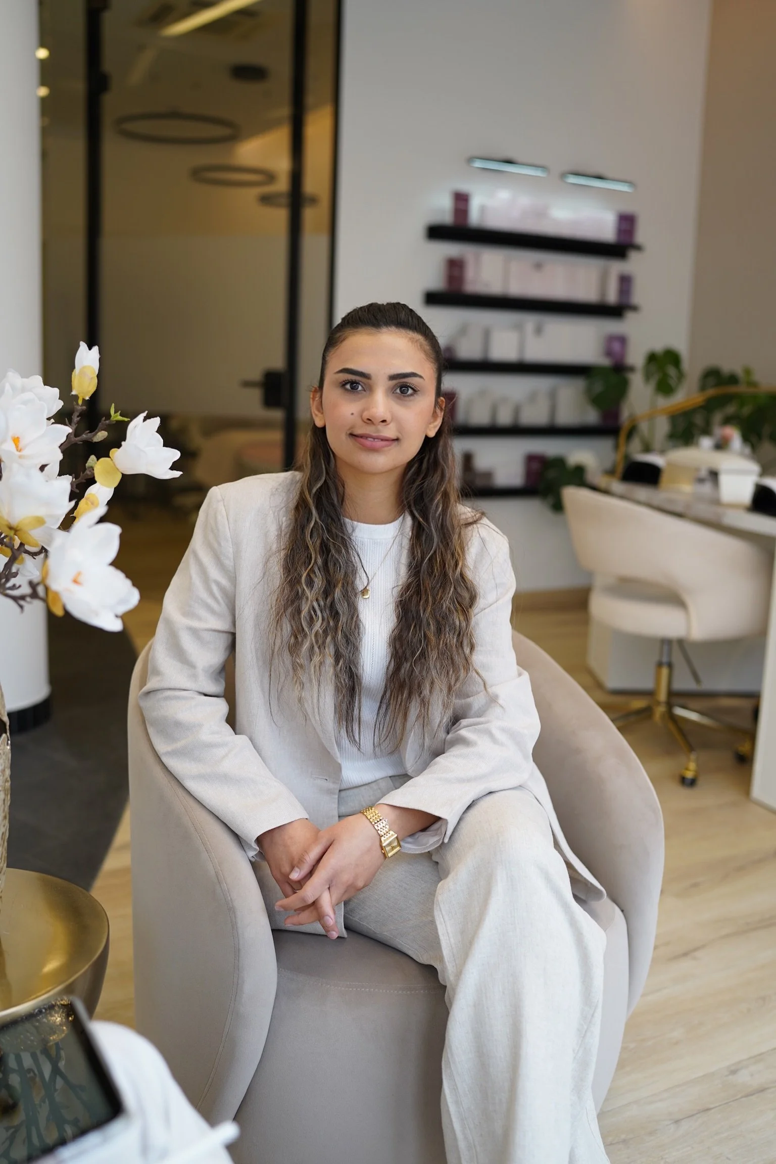 Junge Frau in weißem Anzug sitzt komfortabel auf einem Stuhl in modernem Innenraum, mit Blumen im Vordergrund, durchgehende Holzböden und Regale im Hintergrund.