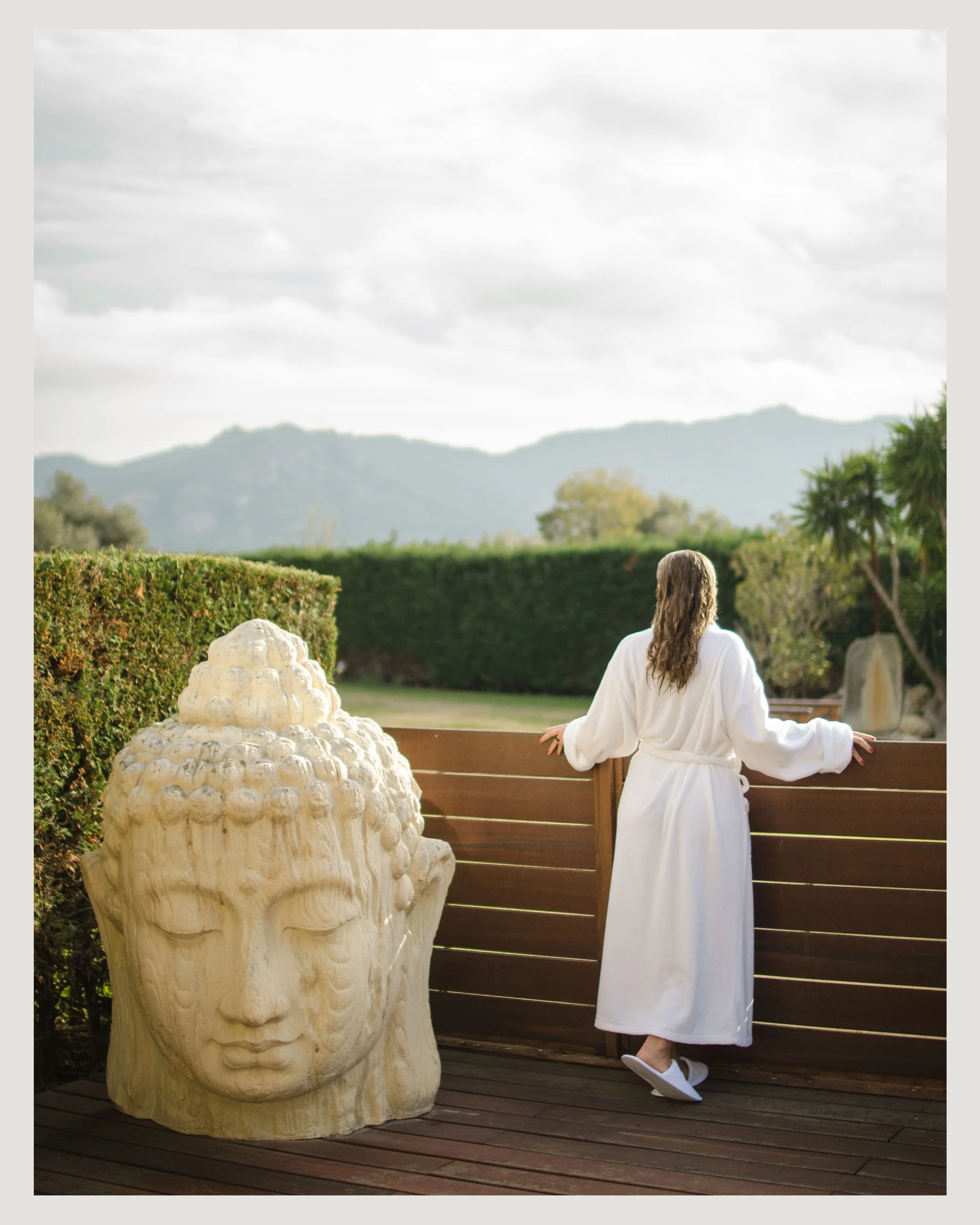Una persona con bata blanca de espaldas, de cabello largo, mirando un jardín con montañas al fondo, rodeada de plantas y con una escultura grande de cabeza de Buda de piedra a la izquierda. En el hotel Mas Tapiolas de la cadena Sallés hotels.