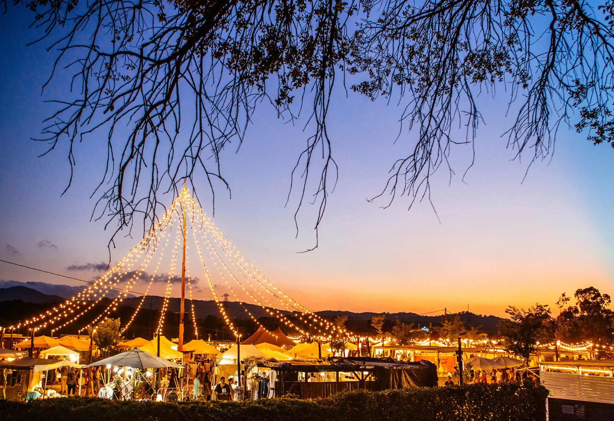 Un mercado nocturno al atardecer con puestos y luces decorativas, árboles en primer plano y montañas al fondo.