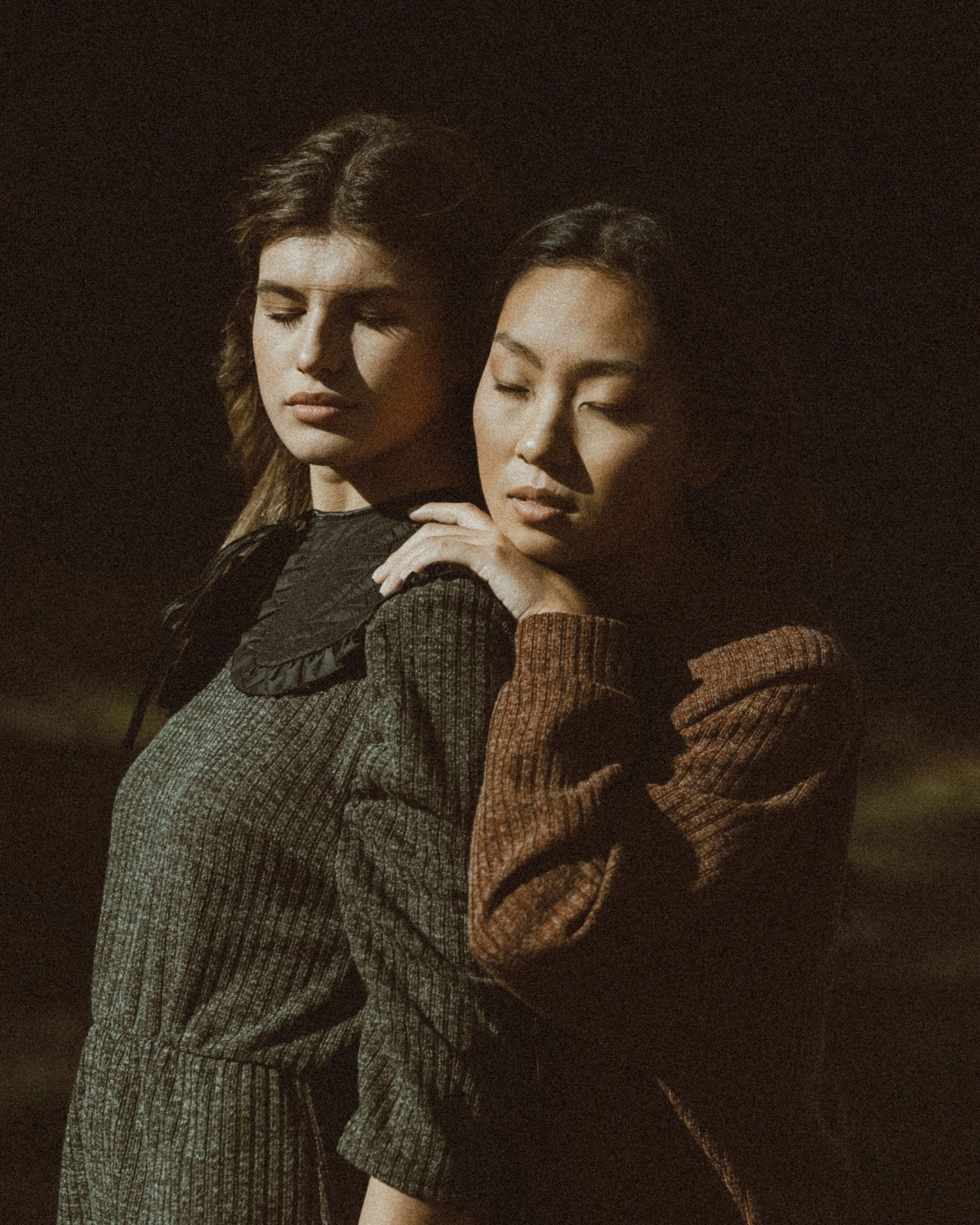 Dos mujeres con los ojos cerrados, una apoyando la cabeza en el hombro de la otra en un ambiente oscuro.