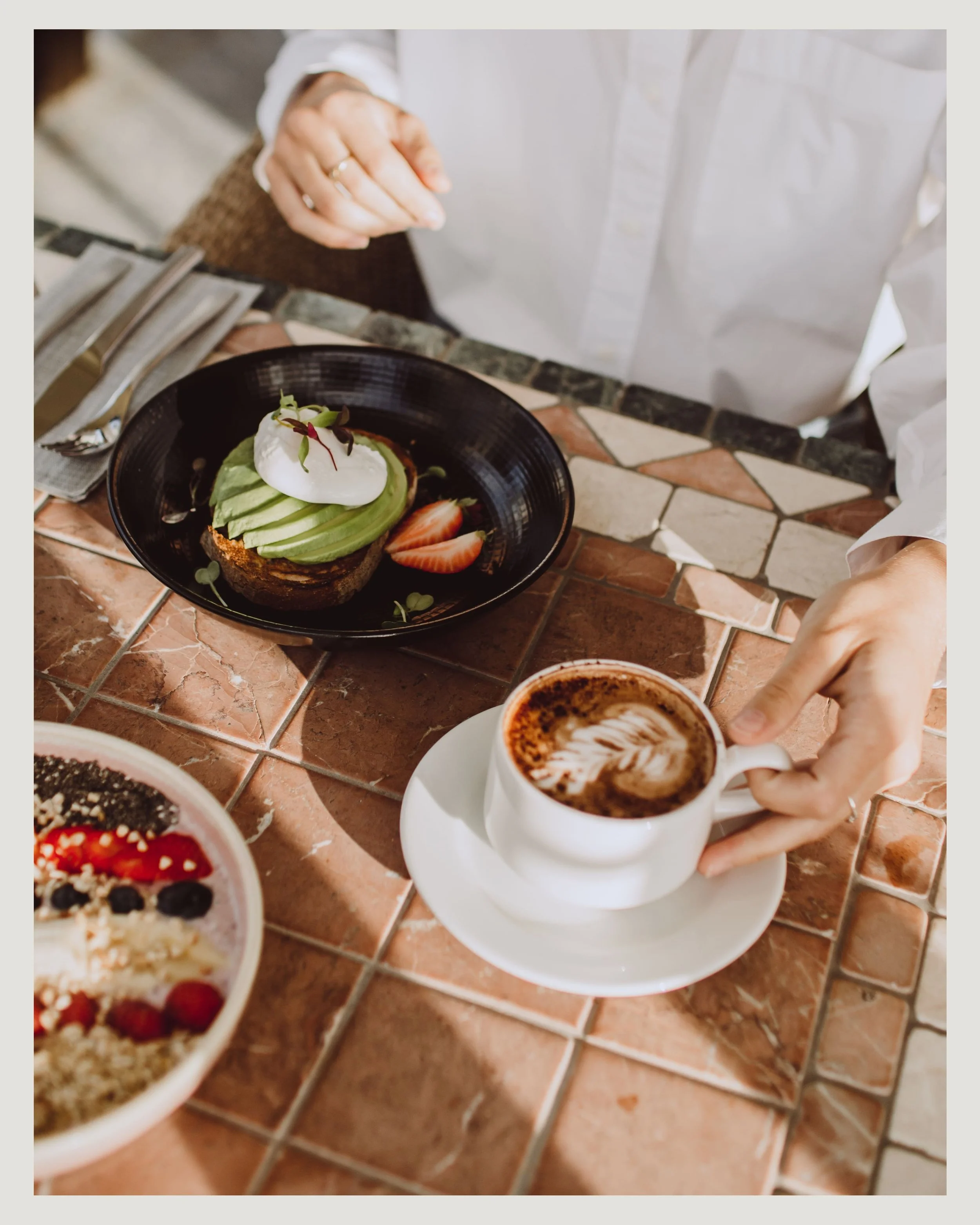 Persona sirviendo una taza de café con arte de leche en una mesa de cerámica, con un plato de aguacate, tomate, huevo y pan, y un bol de fruta con mezcla de berries y granola. En el hotel Cala del Pi de la cadena Sallés hotels.