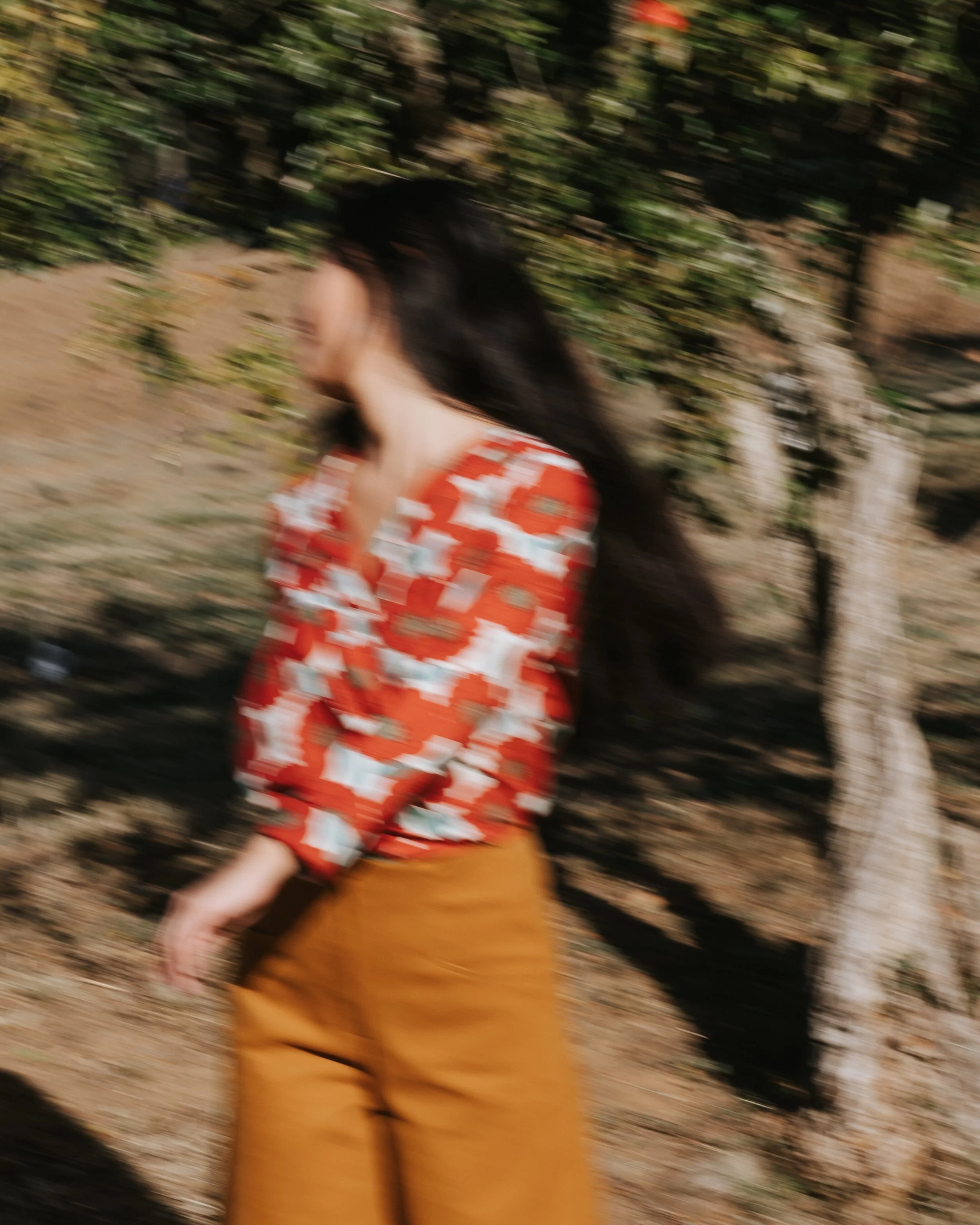 Mujer de cabello largo y oscuro, vestida con blusa roja con patrones blancos, caminando en un campo con árboles. La imagen está borrosa por el movimiento.