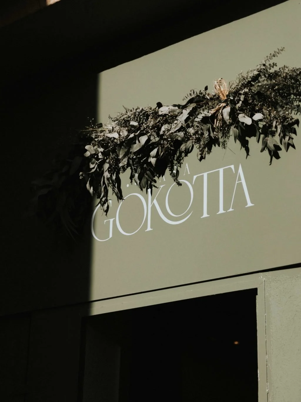 Cartel que dice 'Gatokiota' con decoración de ramas y hojas verdes, en un fondo de pared blanca y sombra