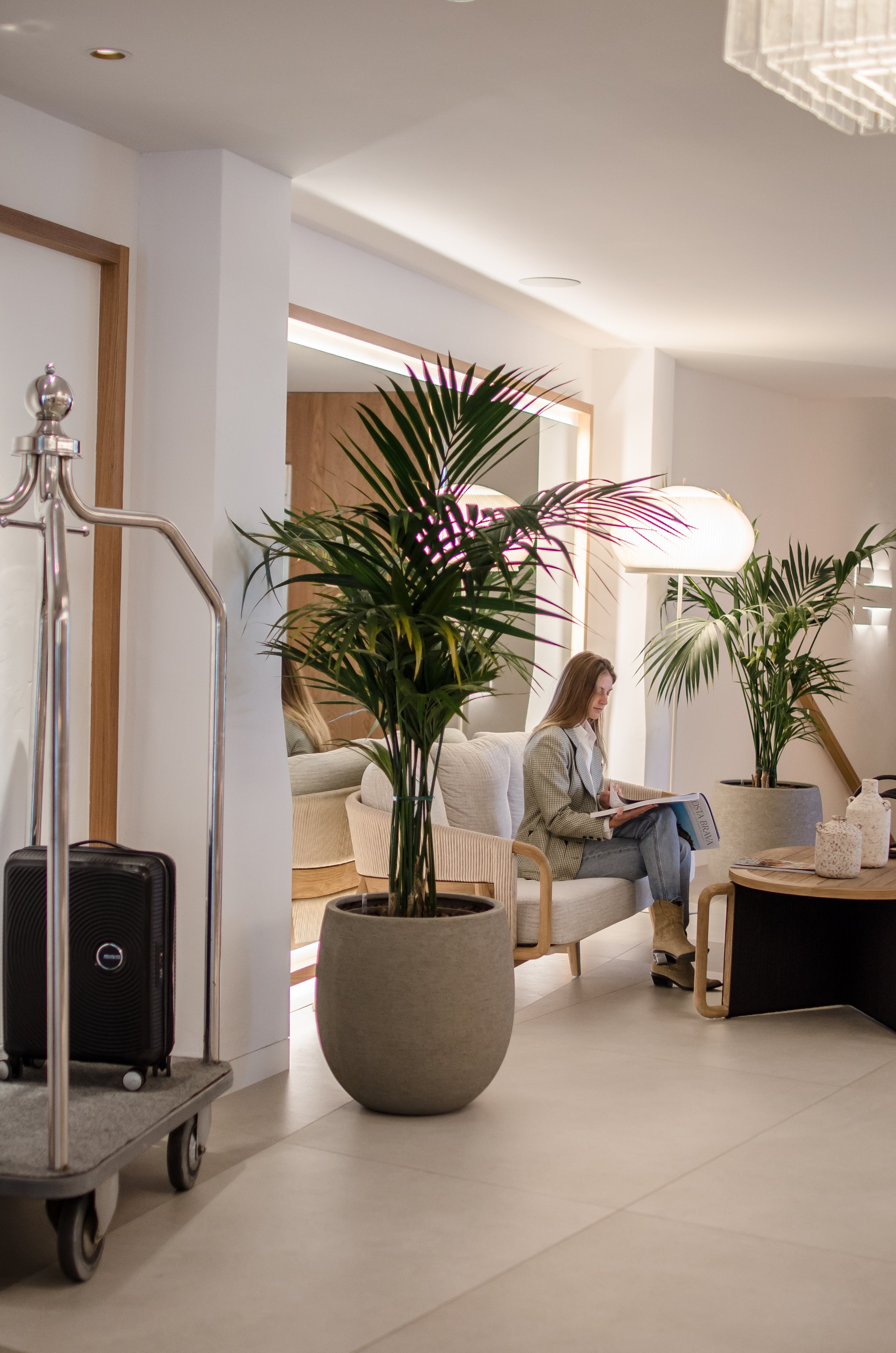 Mujer sentada leyendo un periódico en una sala de hotel con plantas, sillón y lámparas.