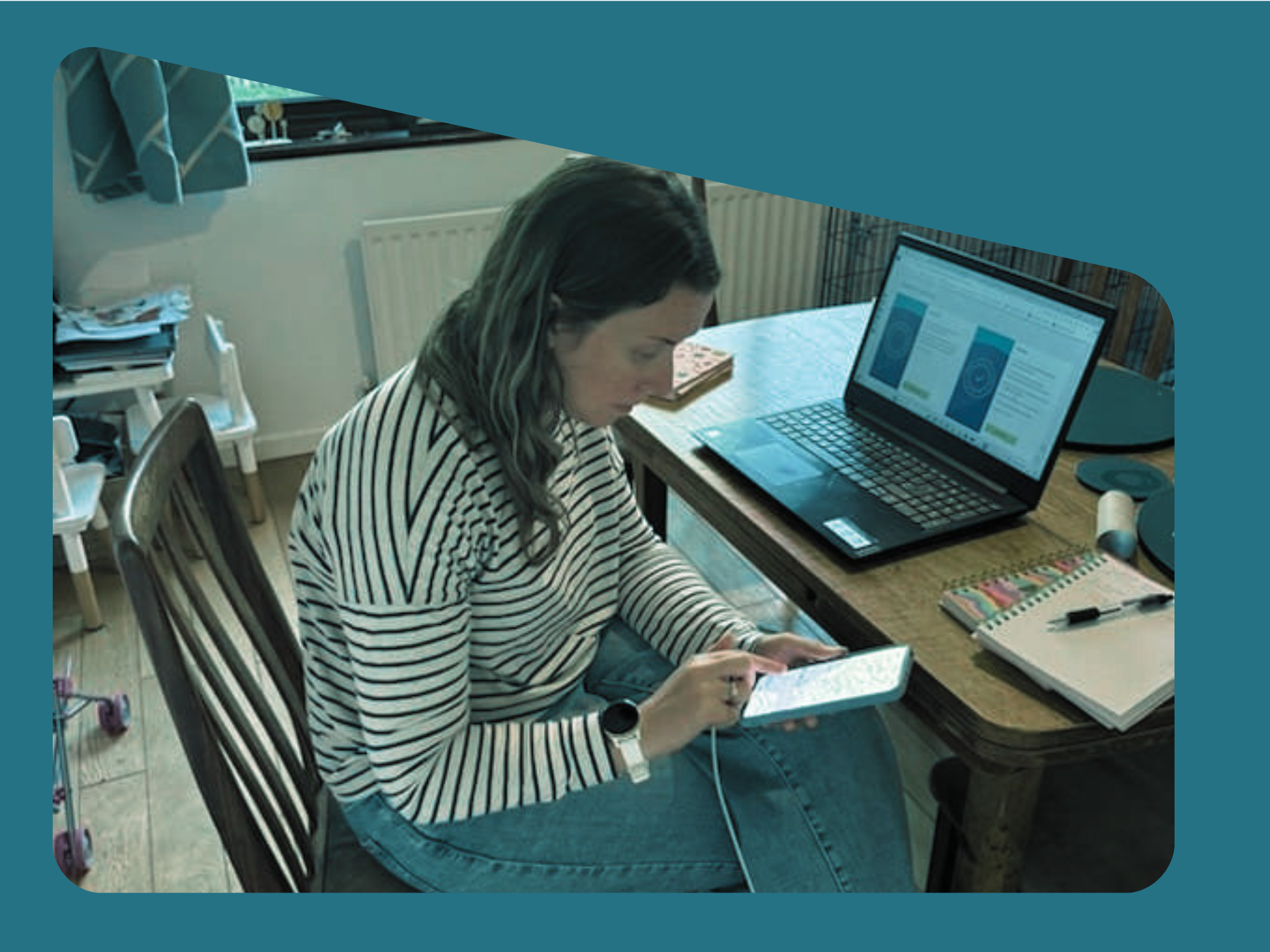 A woman with long dark hair wearing a striped shirt is sitting at a wooden table, holding a tablet in her hands. On the table, there is a laptop displaying charts or graphs, along with some notebooks, pens, and other office supplies. The background shows a room with books, a chair, and a box with papers.