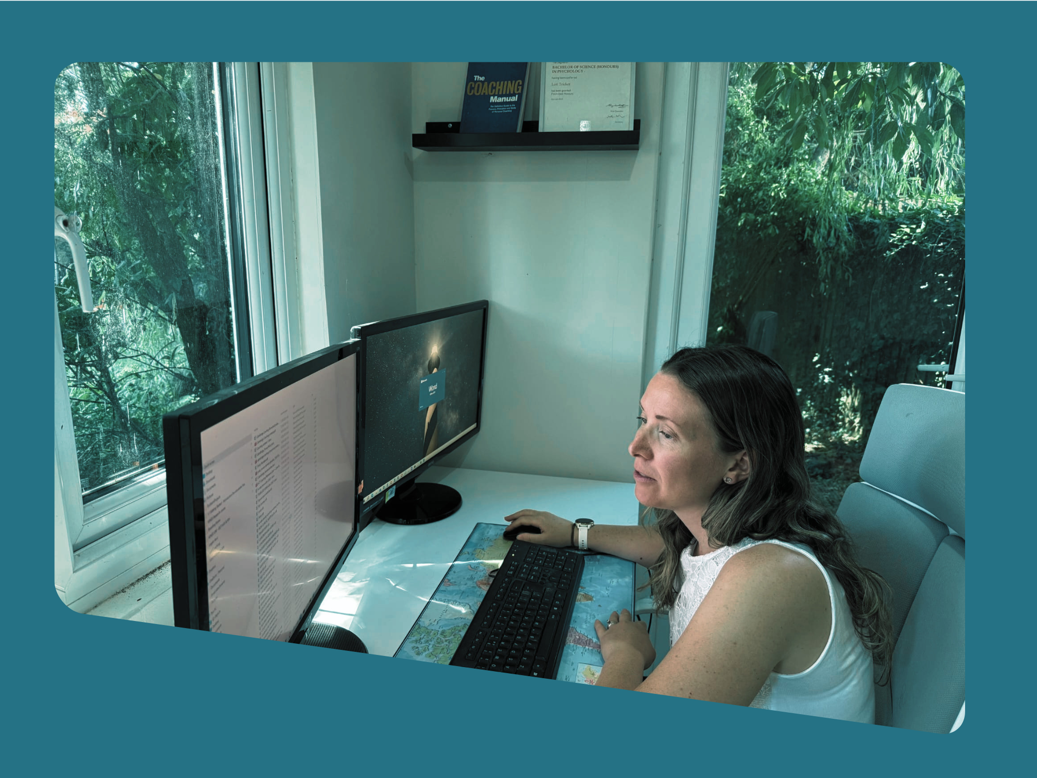 A woman working at a desk in an office with dual computer monitors, a keyboard, and a mouse, while looking at one of the screens. Behind her, large windows reveal green trees outside, and there are books and framed certificates on a shelf on the wall.