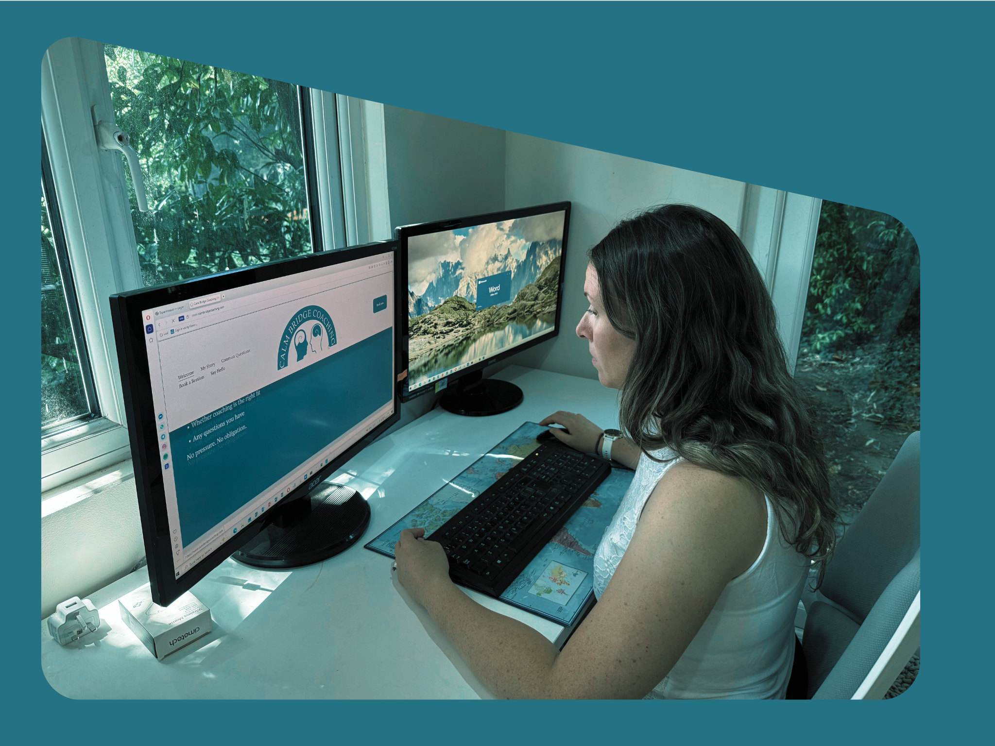 Woman with long dark hair working at a desk with dual computer monitors, window with greenery outside behind her.