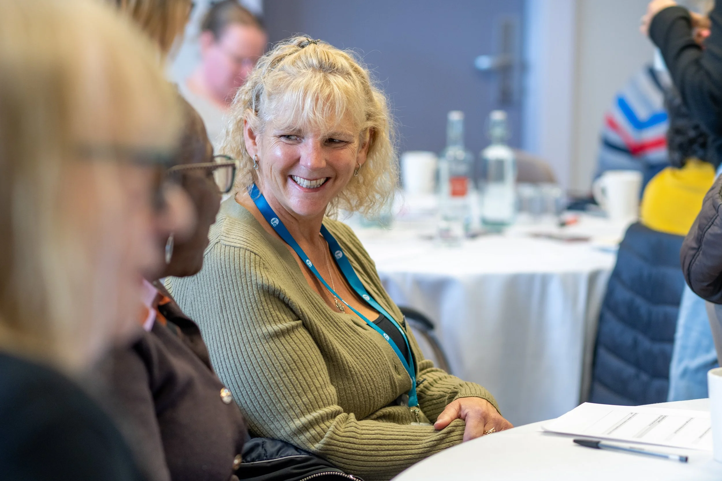 A Swindon Carers Together team member with blonde curly hair is smiling and talking to others at a Carers event, wearing a green cardigan and a blue lanyard, seated at a table with papers and a pen.