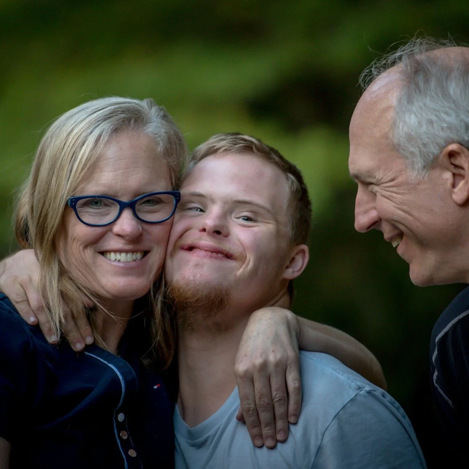 A happy family of three embracing outdoors with green trees in the background.