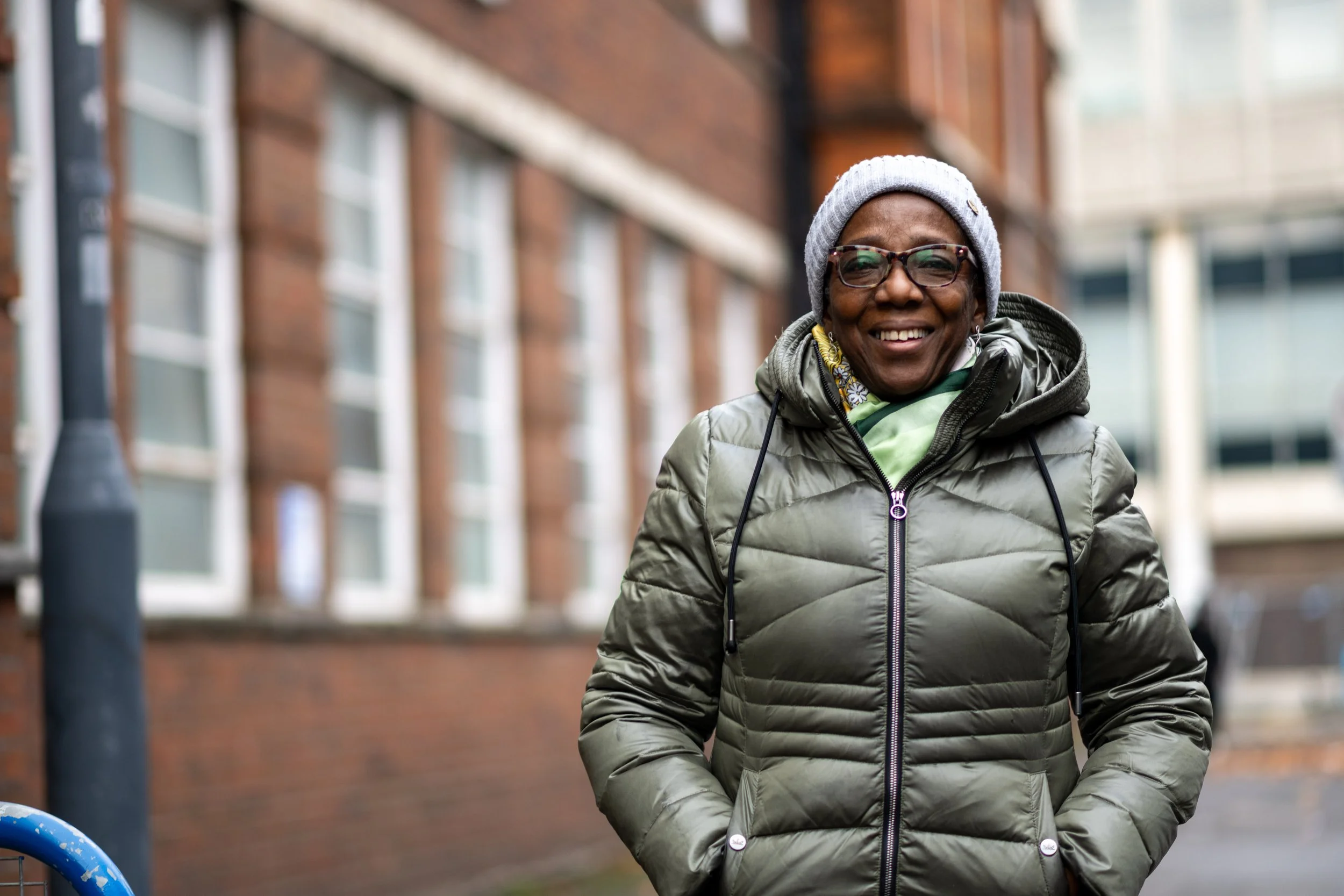 Smiling woman in glasses and a gray beanie walking outdoors, wearing a green jacket with a hoodie layering, outside of Sanford House in Swindon.