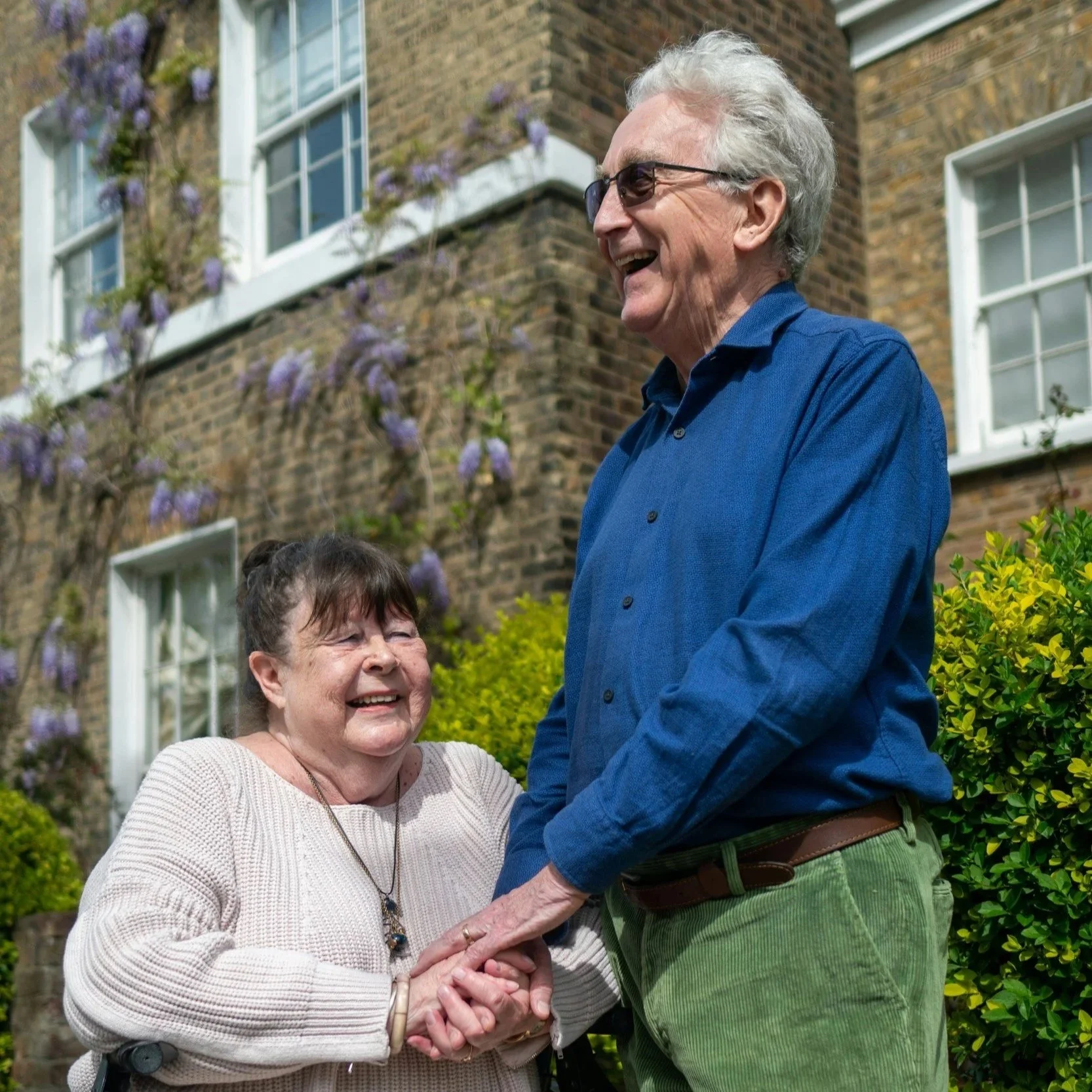 An elderly woman and man standing outside, holding hands, smiling and laughing. The woman is wearing a beige sweater and jewelry, and the man is in a blue shirt and green pants. There are green bushes and a brick building with purple flowers in the background.