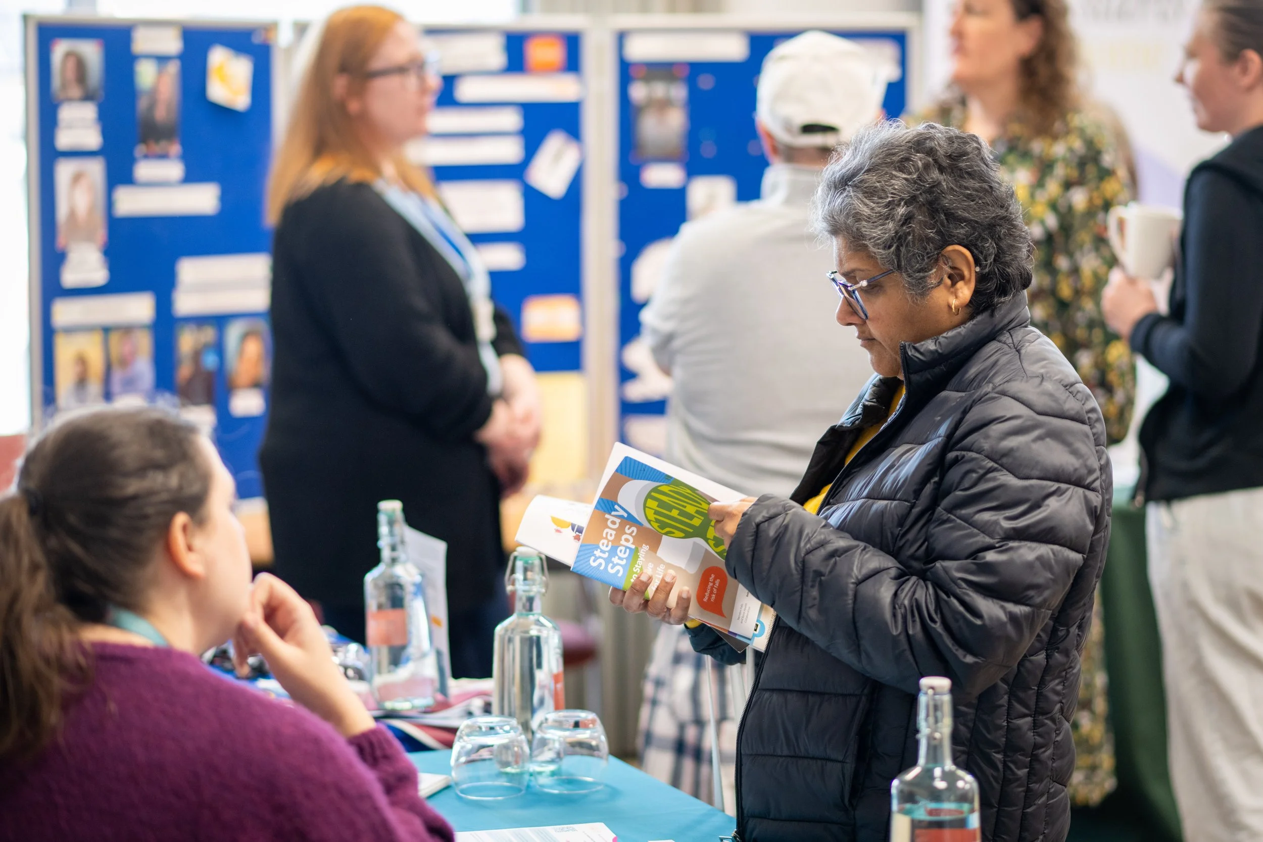 A woman with gray curly hair and glasses reading a brochure at a table with water bottles, surrounded by people at a Carers event.