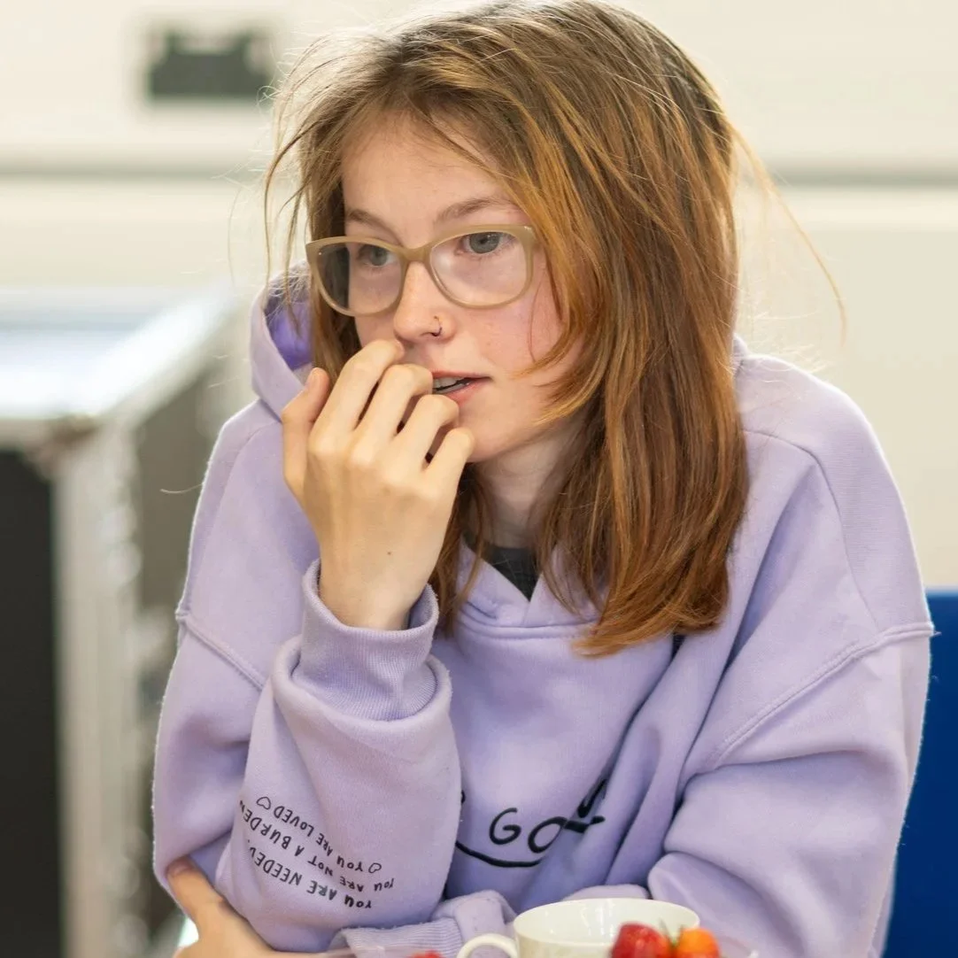 A young woman with red hair, glasses, and a nose piercing, wearing a lavender hoodie, sits with her hand near her mouth in a thoughtful moment, at a table with strawberries in a cup in the foreground.