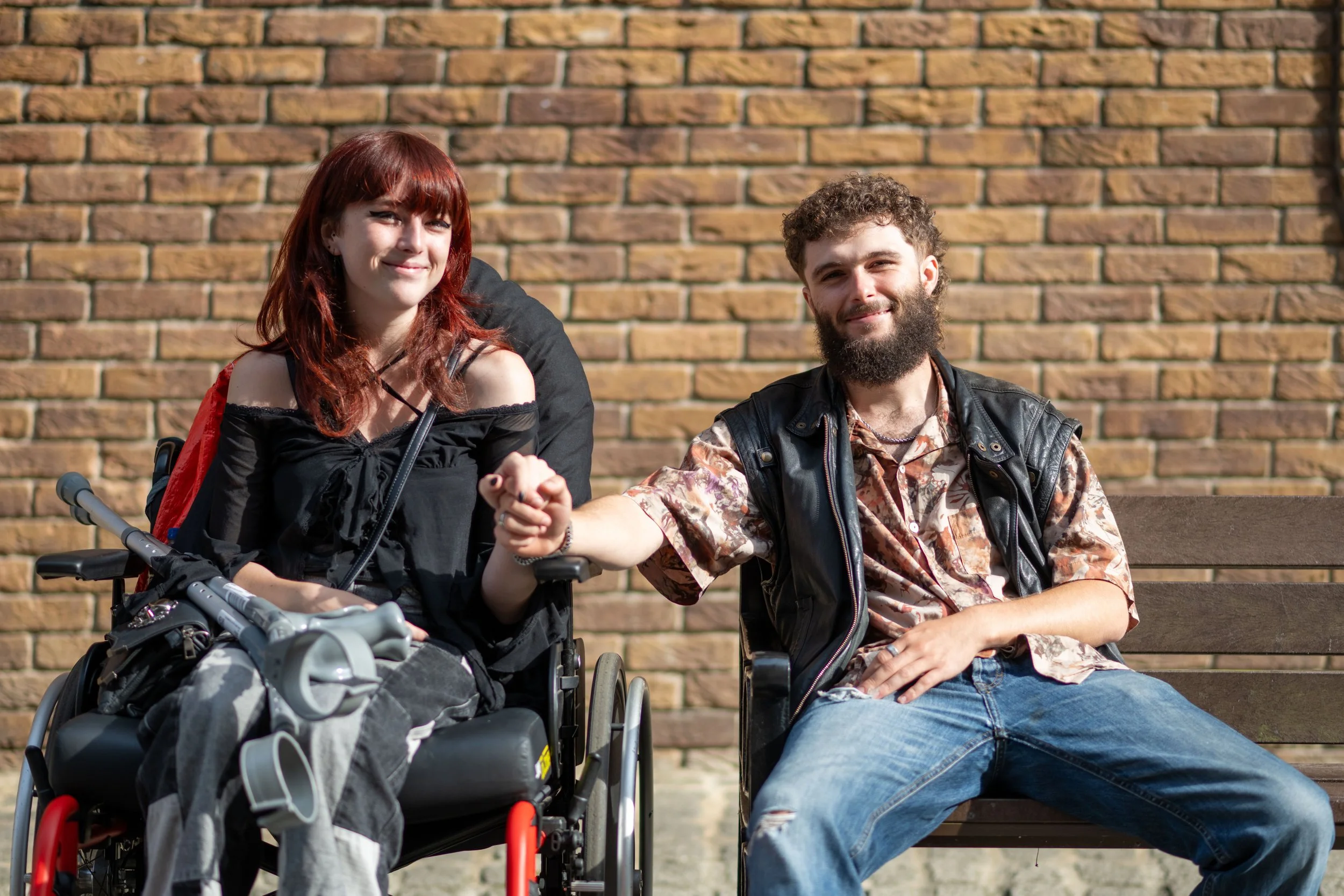 A woman in a wheelchair and a man sitting on a park bench holding hands, both smiling. The woman has red hair and the man has a beard and curly hair. They are sitting in front of a brick wall.