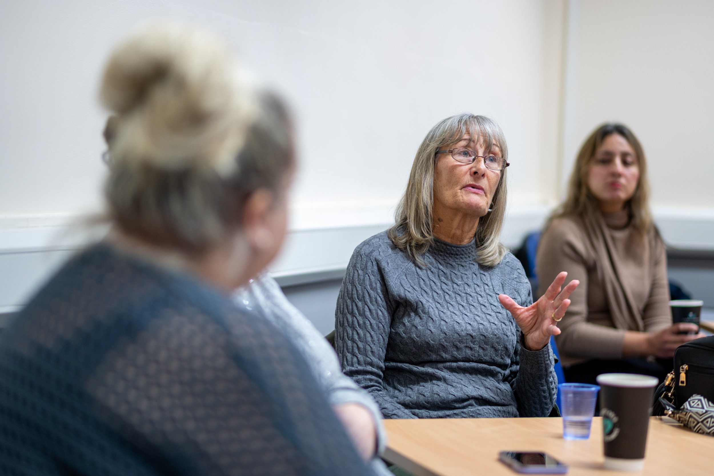 A woman with gray hair, glasses, and a gray sweater speaking during a meeting with three other women seated at a table.